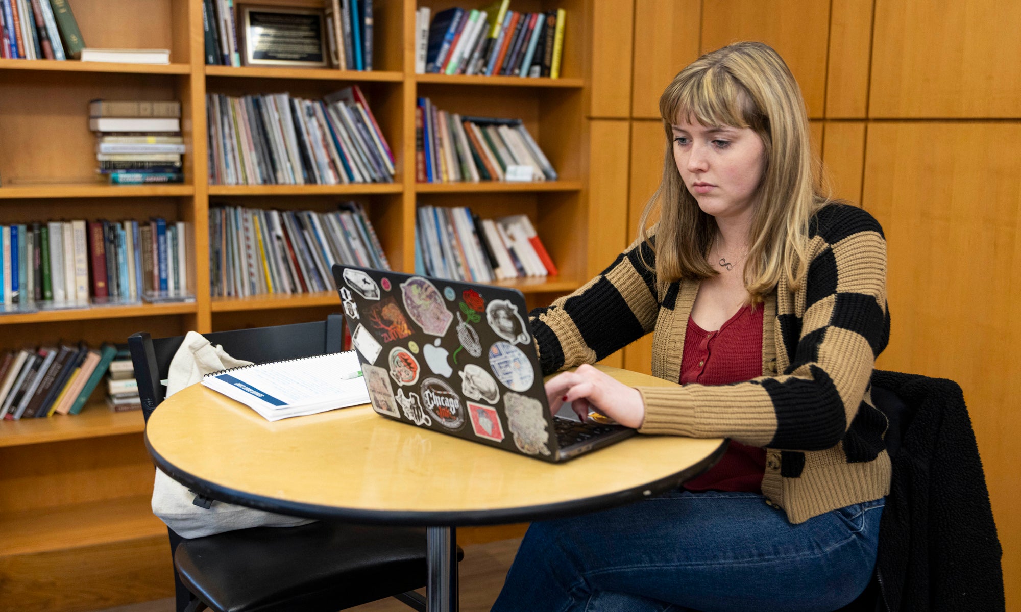 A woman with a laptop sitting at a small table in front of a bookcase.