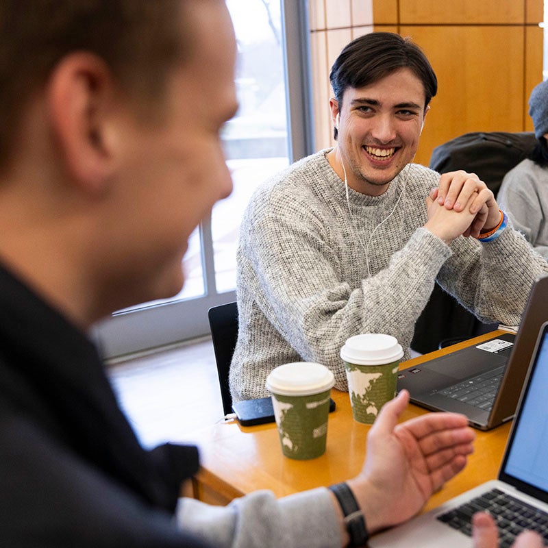 Two people sit at a table with laptops and coffee cups, engaged in conversation.