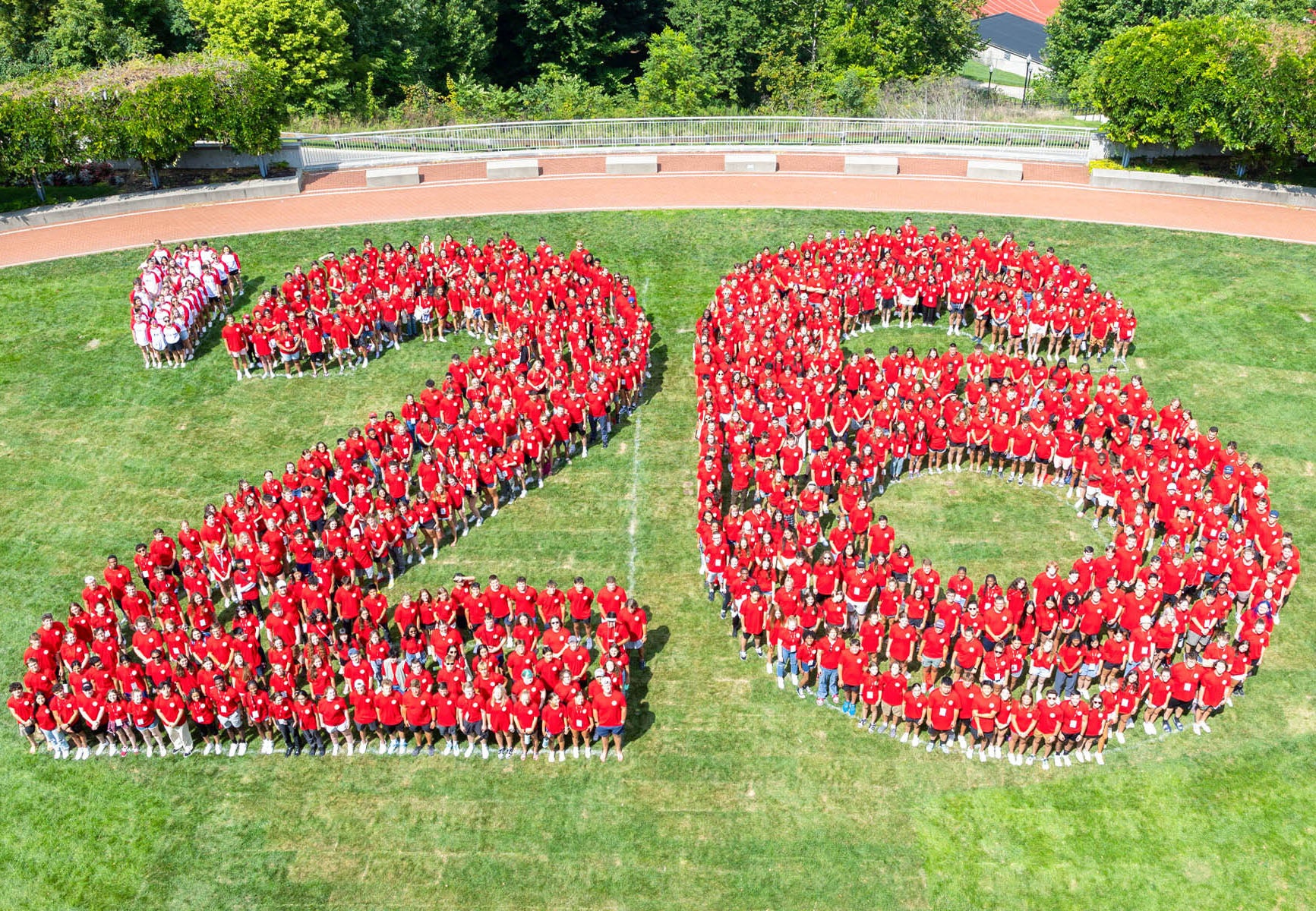 Aerial view of hundreds of people in red tee shirts standing on a large grassy oval, grouped into a block formation of the number 26.