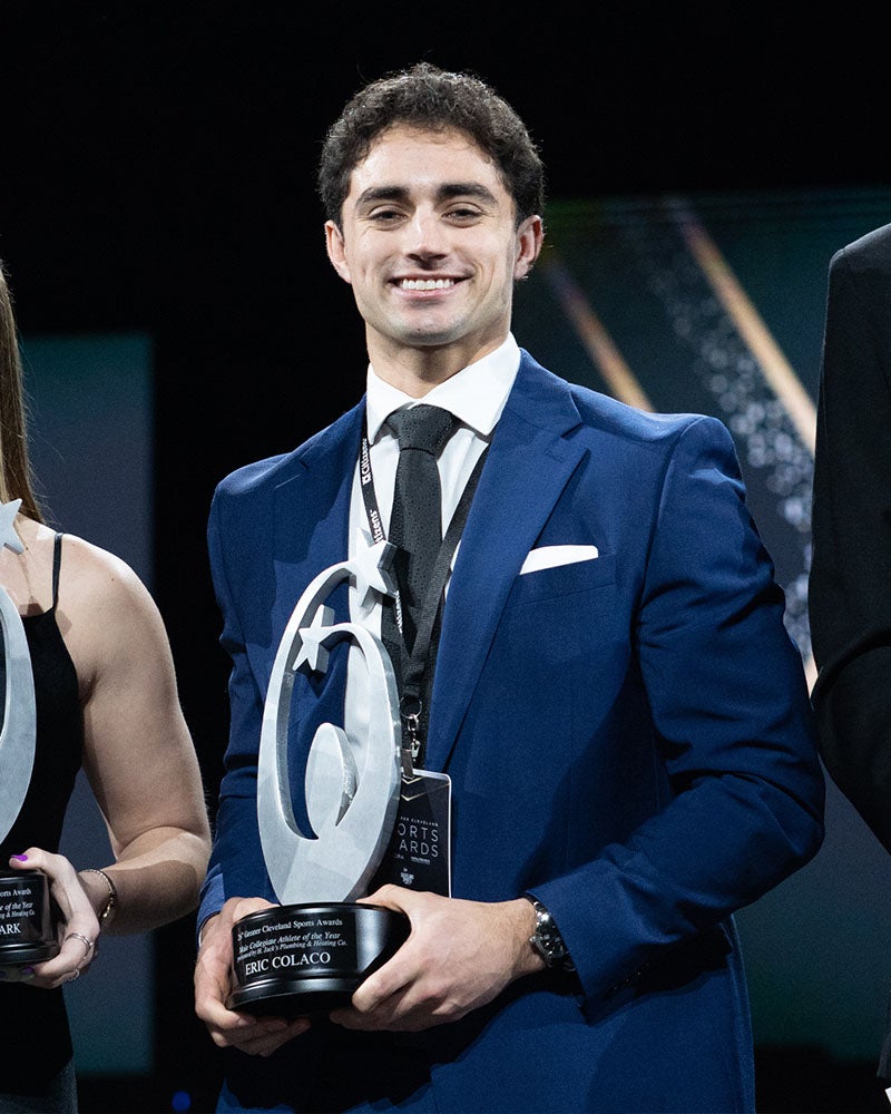Photo of Denison alum Eric Colaco ’25 in a blue suit holding a trophy award.