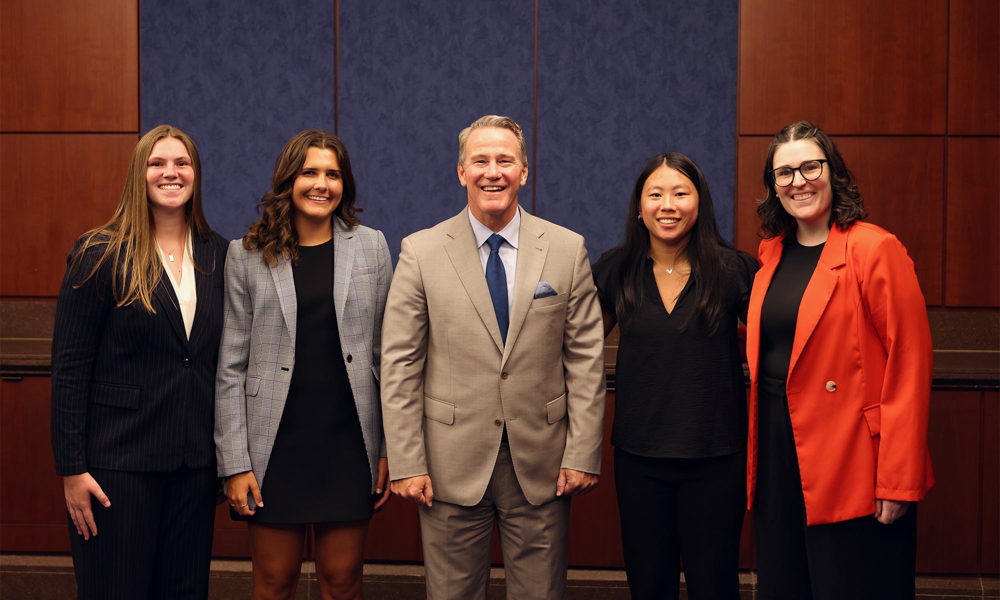 Five people standing side by side, dressed in business attire, posing for a group photo in front of a blue and brown wall.