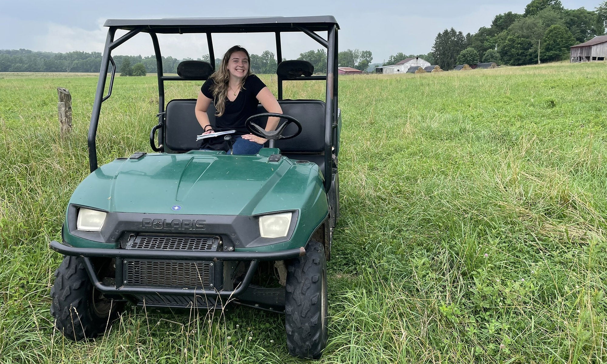 A Denison student sits in the driver’s seat of a green utility vehicle parked on grass, with a field and trees in the background under a cloudy sky.