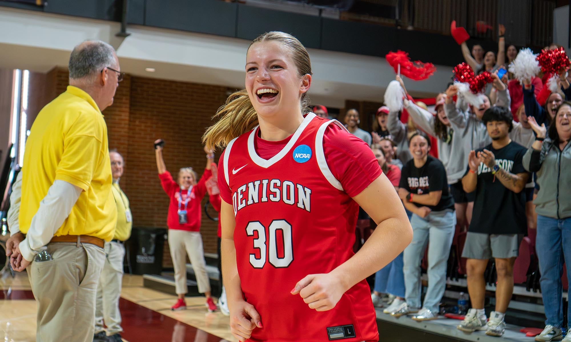 A basketball player wearing a red Denison jersey with the number 30 smiles while walking on a court. A man in a yellow shirt stands nearby and fans in the background cheer with red and white pom-poms.