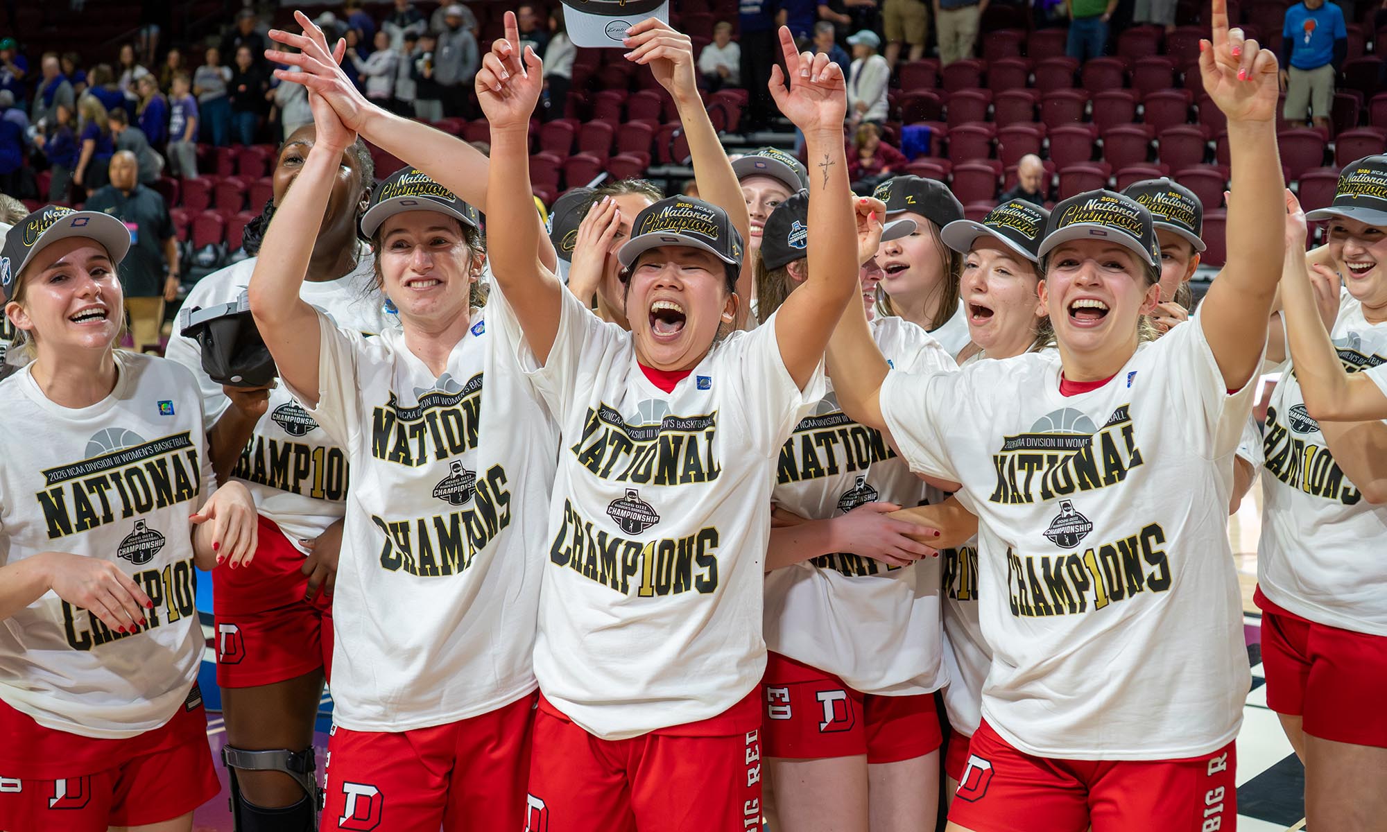 A group of young women in white National Champions shirts and red shorts celebrate with their arms raised after Denison women’s basketball clinched the NCAA title.