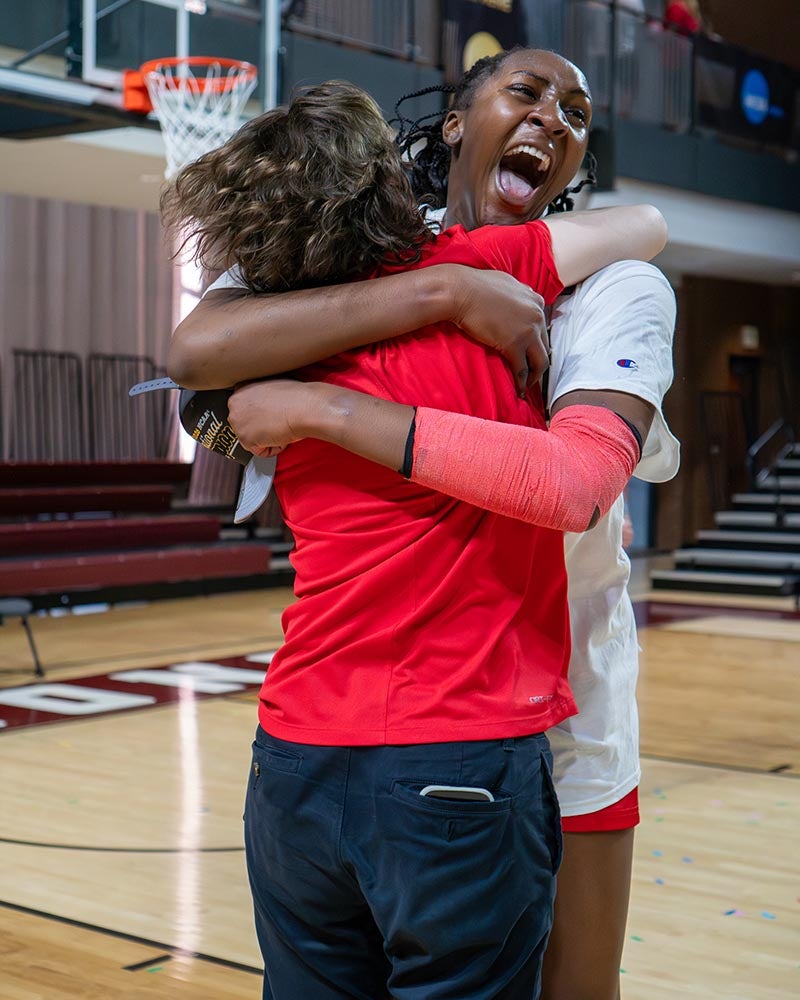 Two people embrace in a gymnasium. One, in athletic shorts and a white shirt, is joyfully shouting—celebrating a Denison women’s basketball victory. The other, wearing a red shirt, has their back to the camera amid the excitement of hoops glory.