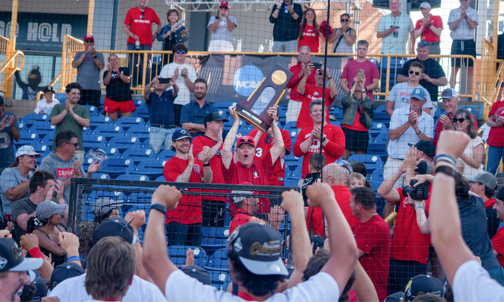 A group of athletes and supporters celebrate on a field, with two people holding up a trophy. Spectators in the background take photos and applaud, and many wear red shirts.