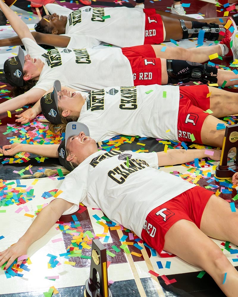 Four Denison women’s basketball players lie on the floor covered in confetti, wearing white t-shirts and red shorts, celebrating their NCAA title win in hoops.