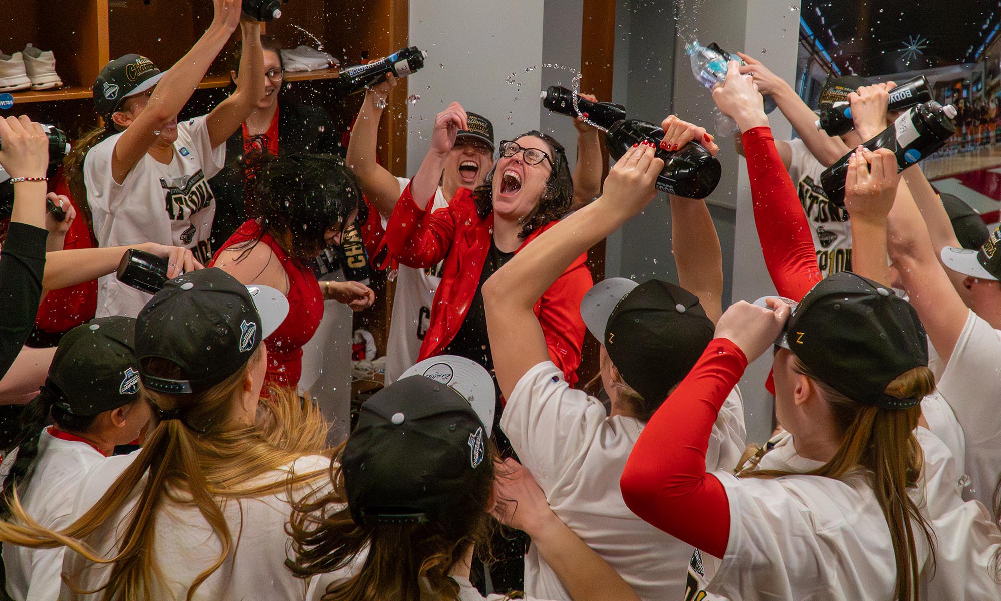 A group of people in matching shirts and hats celebrate indoors, spraying drinks and cheering, as Denison women’s basketball clinches the NCAA title; one person in a red jacket stands in the center, mouth open in excitement.