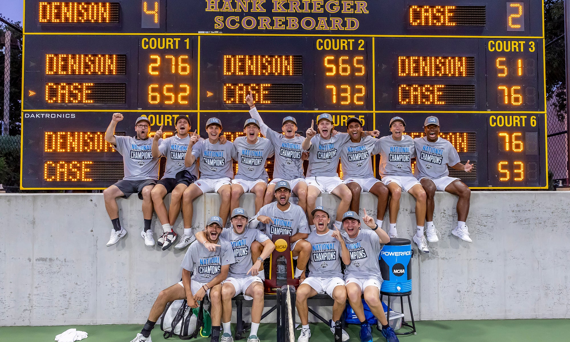 A men's tennis team in matching "National Champion" shirts poses in front of a scoreboard, celebrating their victory with medals and a trophy.
