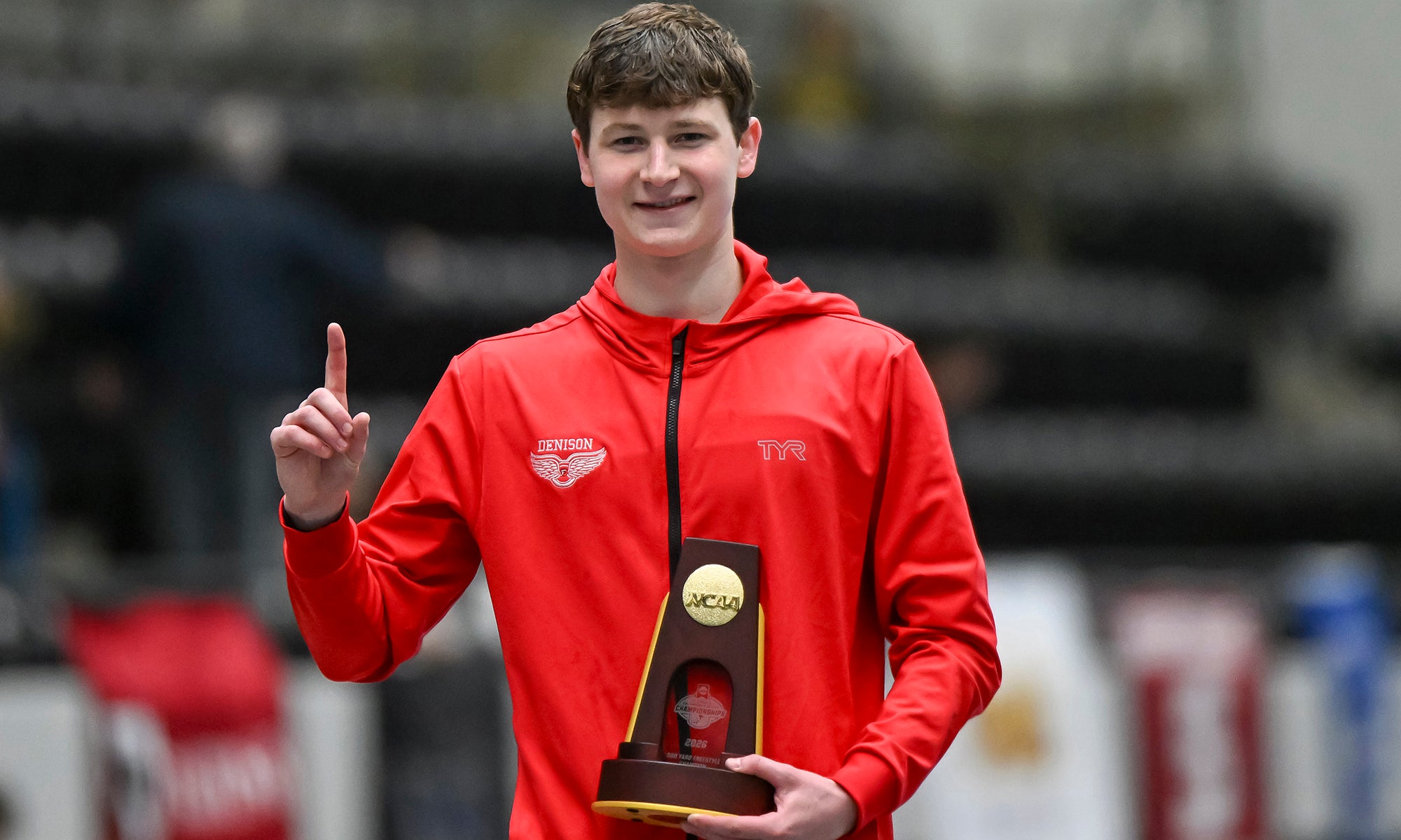 A student athlete in a red hoodie holds a championship trophy in one hand and smile for the camera while making a "number one" sign with his other hand.
