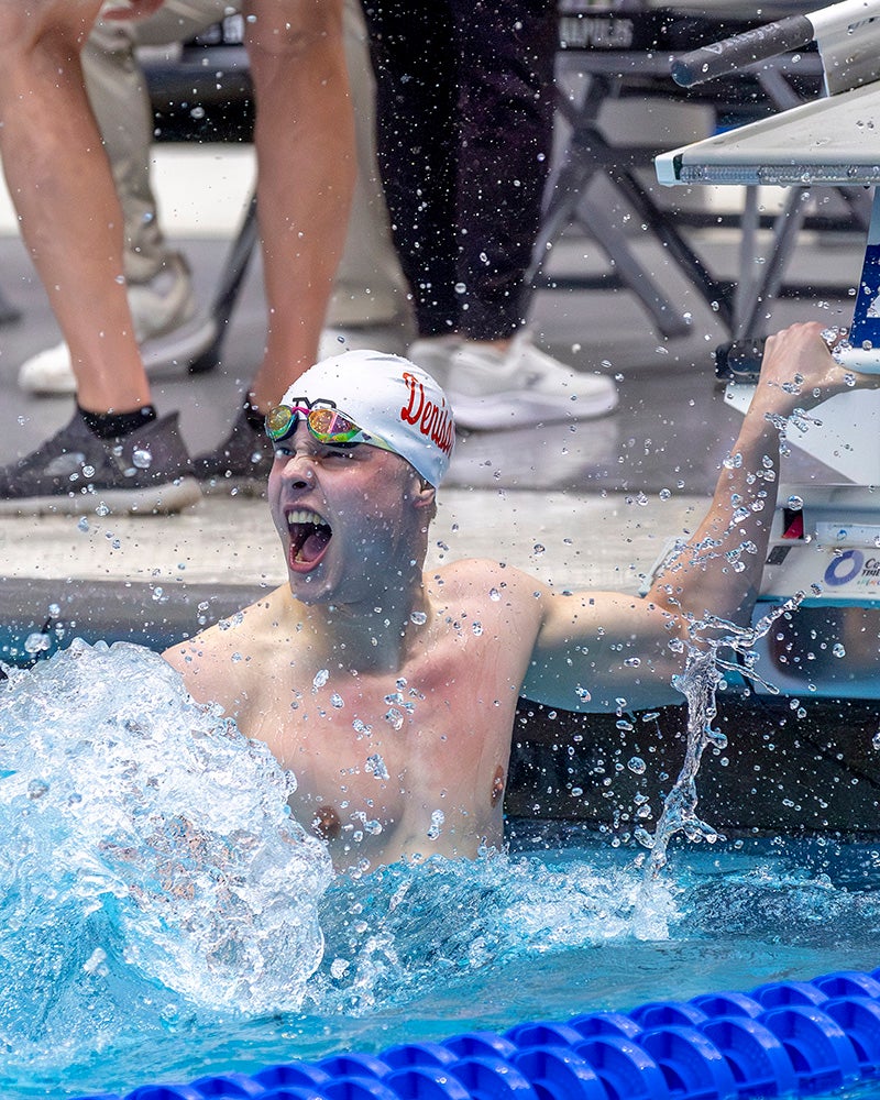 A swimmer wearing goggles and a swim cap is in a pool, splashing water and shouting with an excited expression. People with sneakers are seen in the background near the poolside.