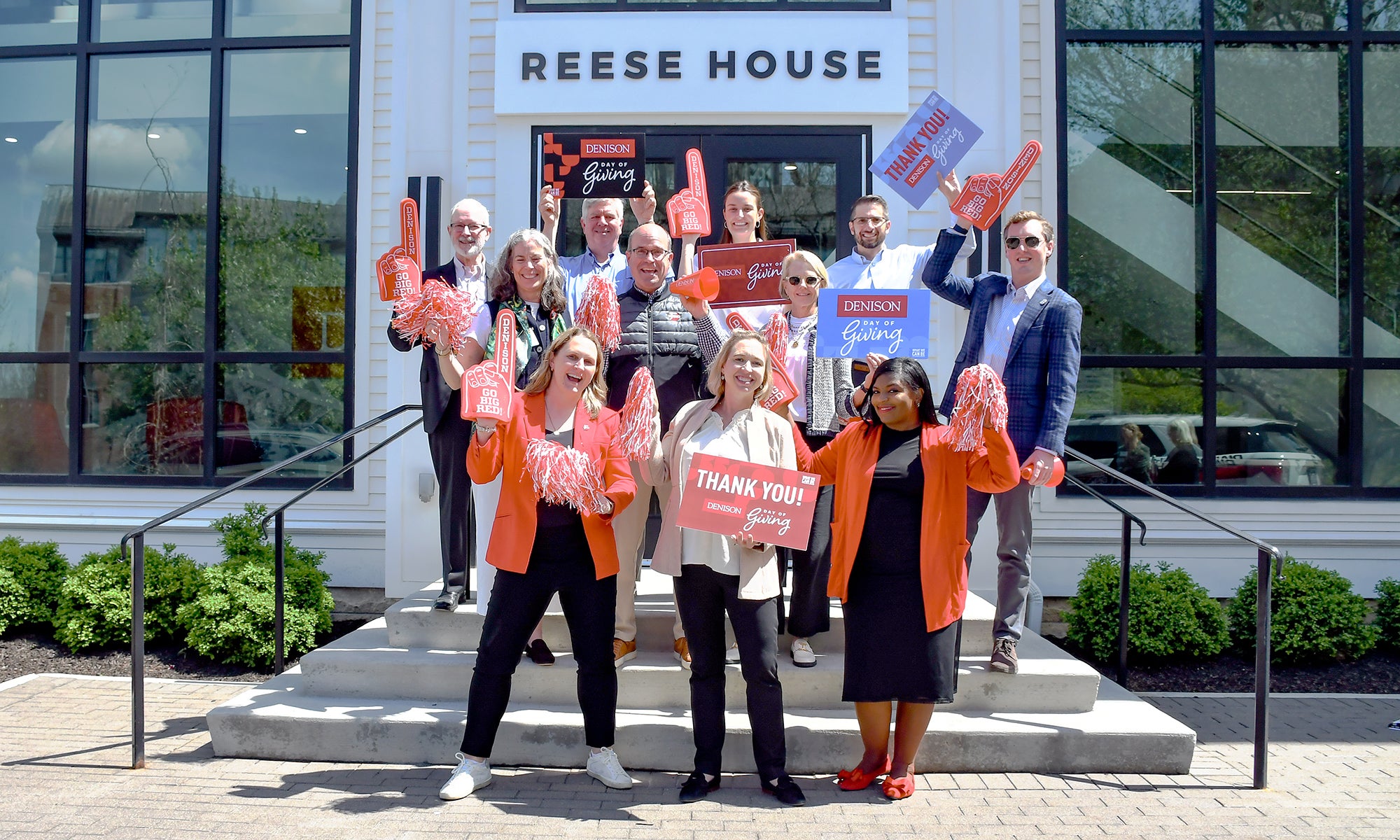 A group of people stand on the steps of a building labeled Reese House, holding signs and pom-poms. Most are wearing orange or holding orange items, and they are posing for a photo in daylight.