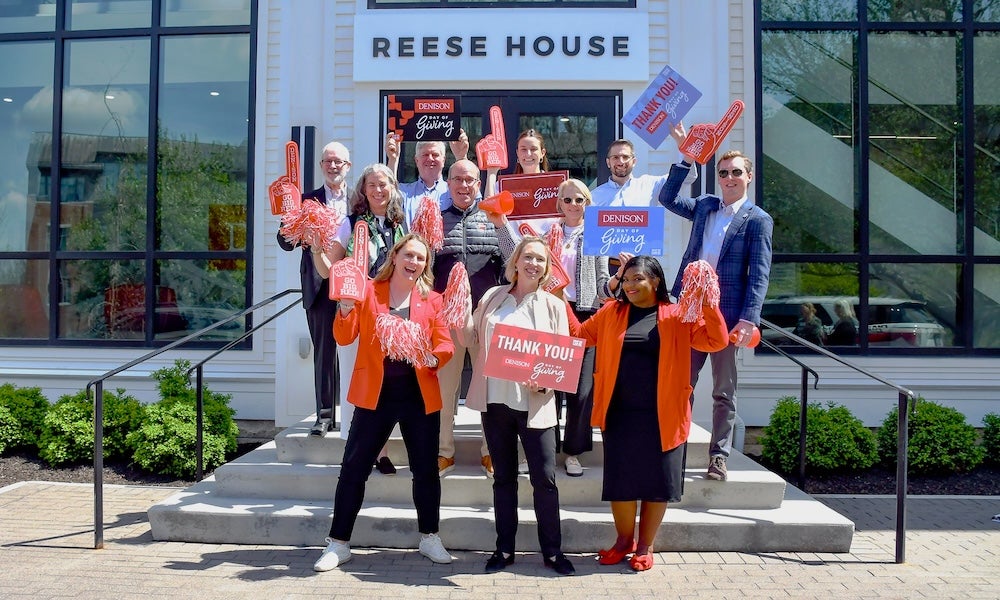 A group of people stand on the steps of a building labeled Reese House, holding signs and pom-poms. Most are wearing orange or holding orange items, and they are posing for a photo in daylight.