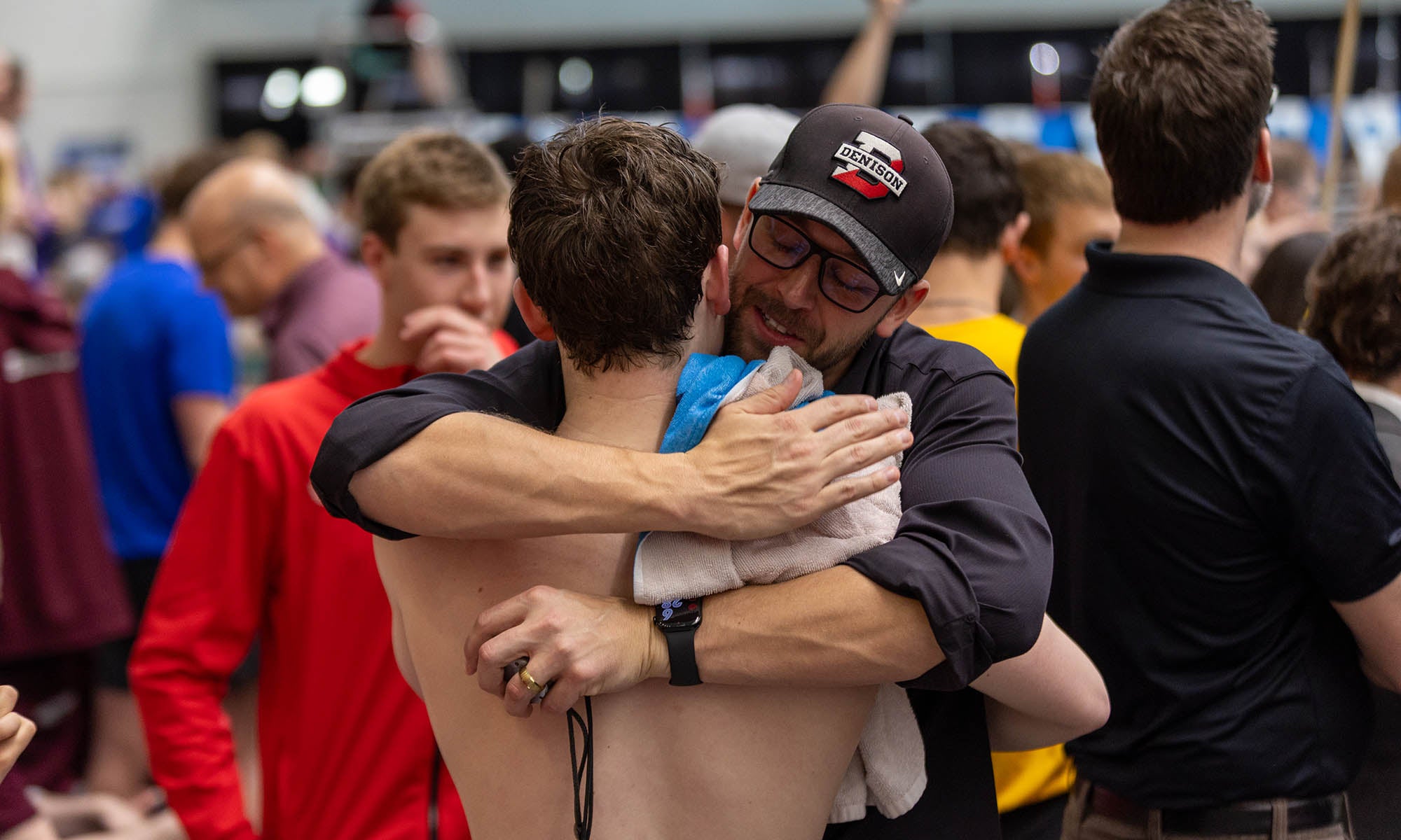 A man in a ball cap hugs a swimmer in the middle of a crowd poolside.