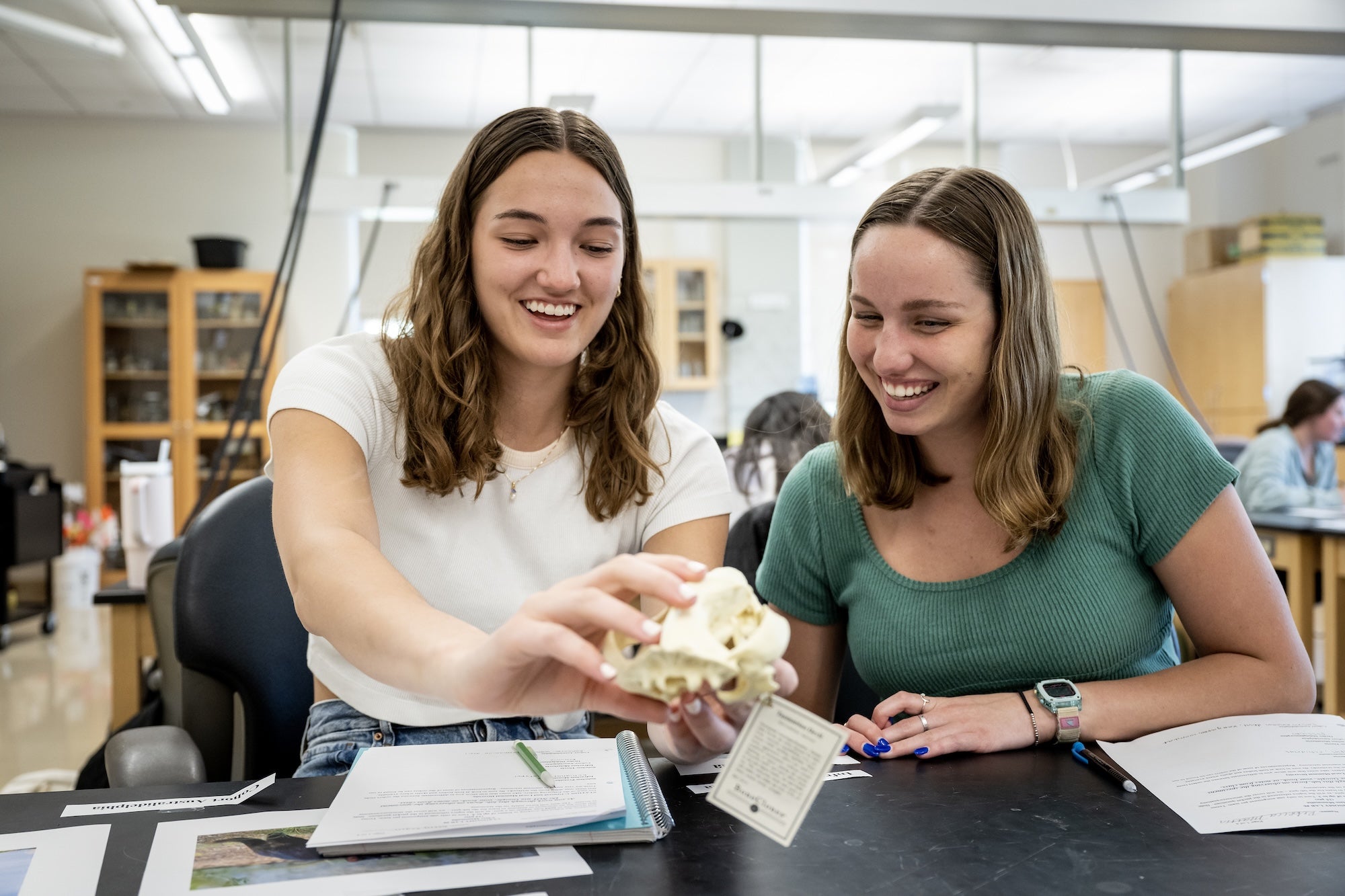 Two smiling women sitting at a lab table examining an animal skull