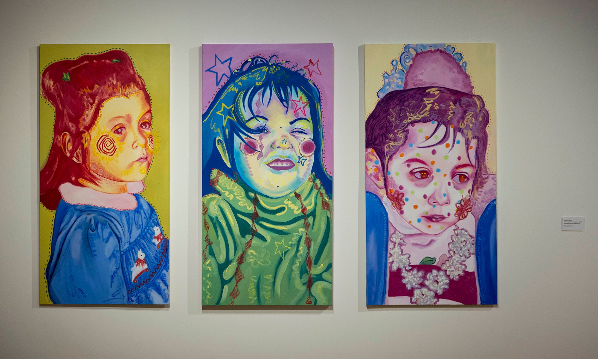 Three colorful portrait paintings hang on a white wall at Denison Museum. Each child, depicted in "It Was Never Just Then," features distinctive facial marks and a unique color palette: yellow-red, green-blue, and pink-purple tones.