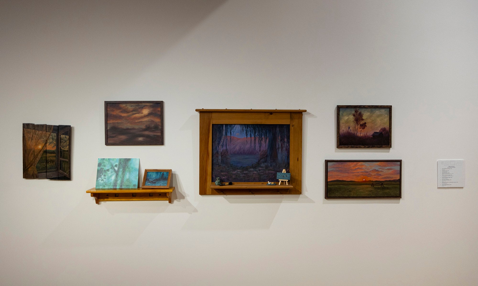 A white wall at the Denison Museum displays seven framed paintings of various sizes for the art exhibition "It Was Never Just Then." One painting sits on a wooden shelf, flanked by two smaller pieces, with a small plaque mounted on the right.