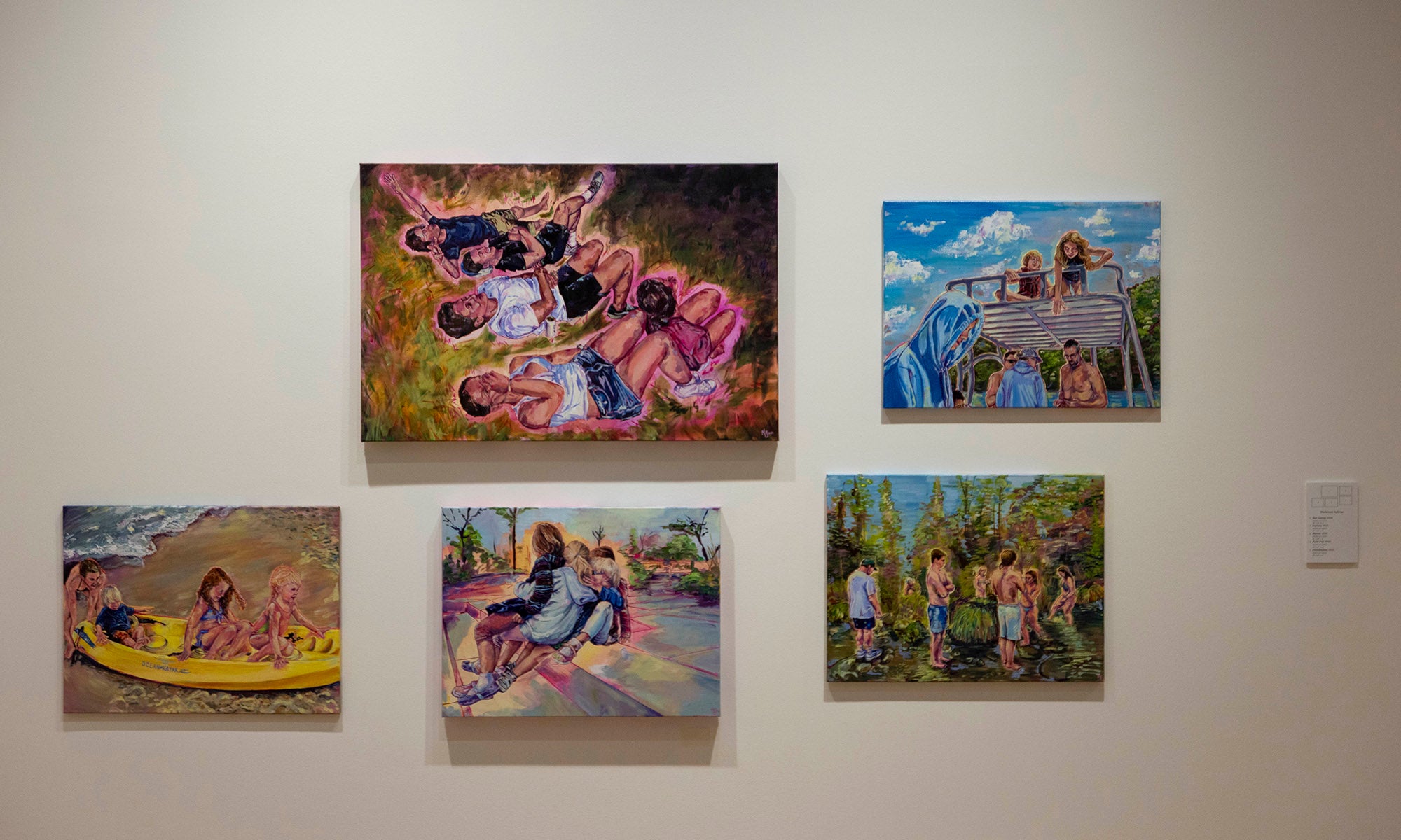 Five colorful paintings are displayed on a white wall at the Denison Museum art exhibition, "It Was Never Just Then"; each features groups of people, some outdoors in nature, engaging in activities like sitting, rowing a boat, and gathering together.