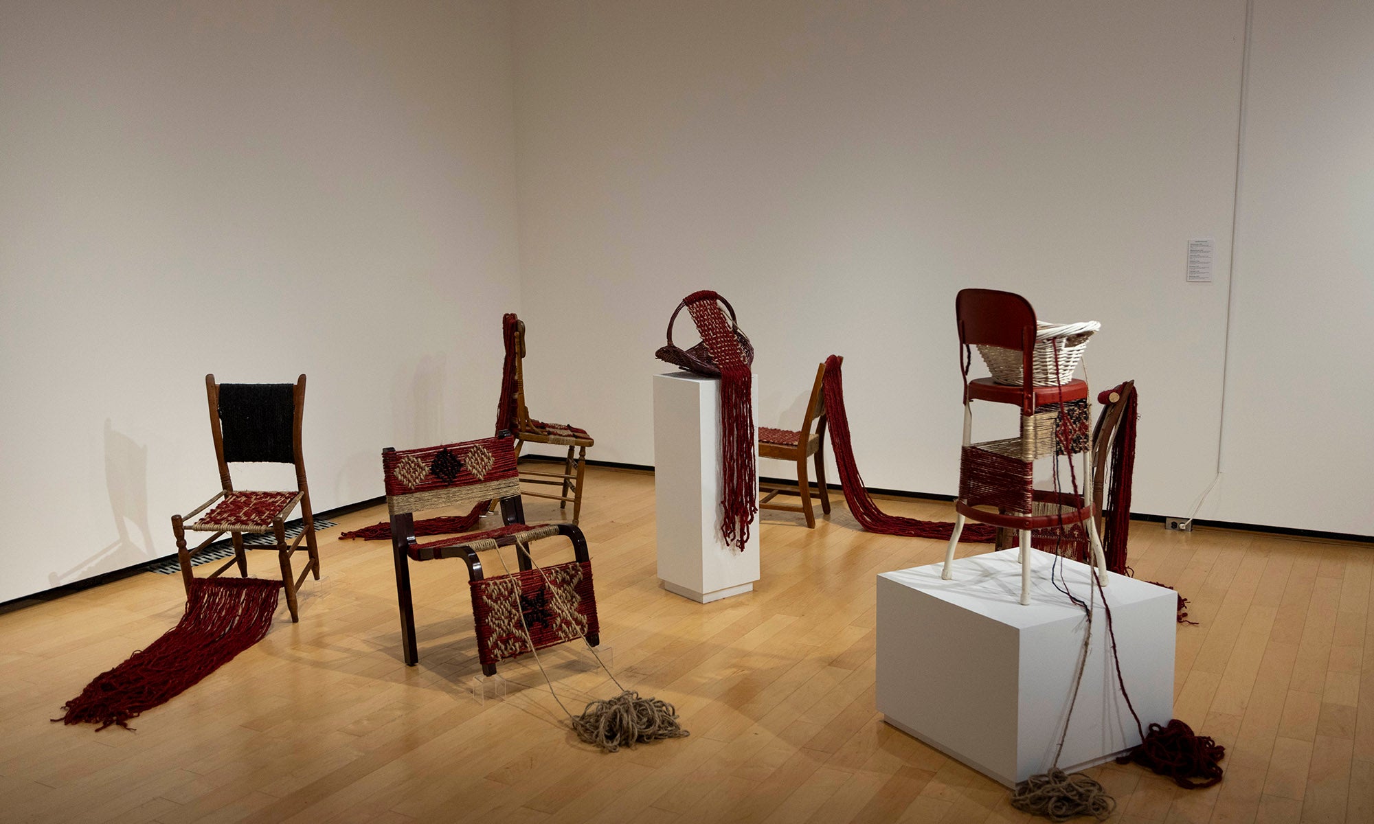 A gallery room at Denison Museum showcases the art exhibition "It Was Never Just Then," with chairs and pedestals draped in woven red and brown scarves. Bundles of yarn rest on the wooden floor, set against pristine white walls.