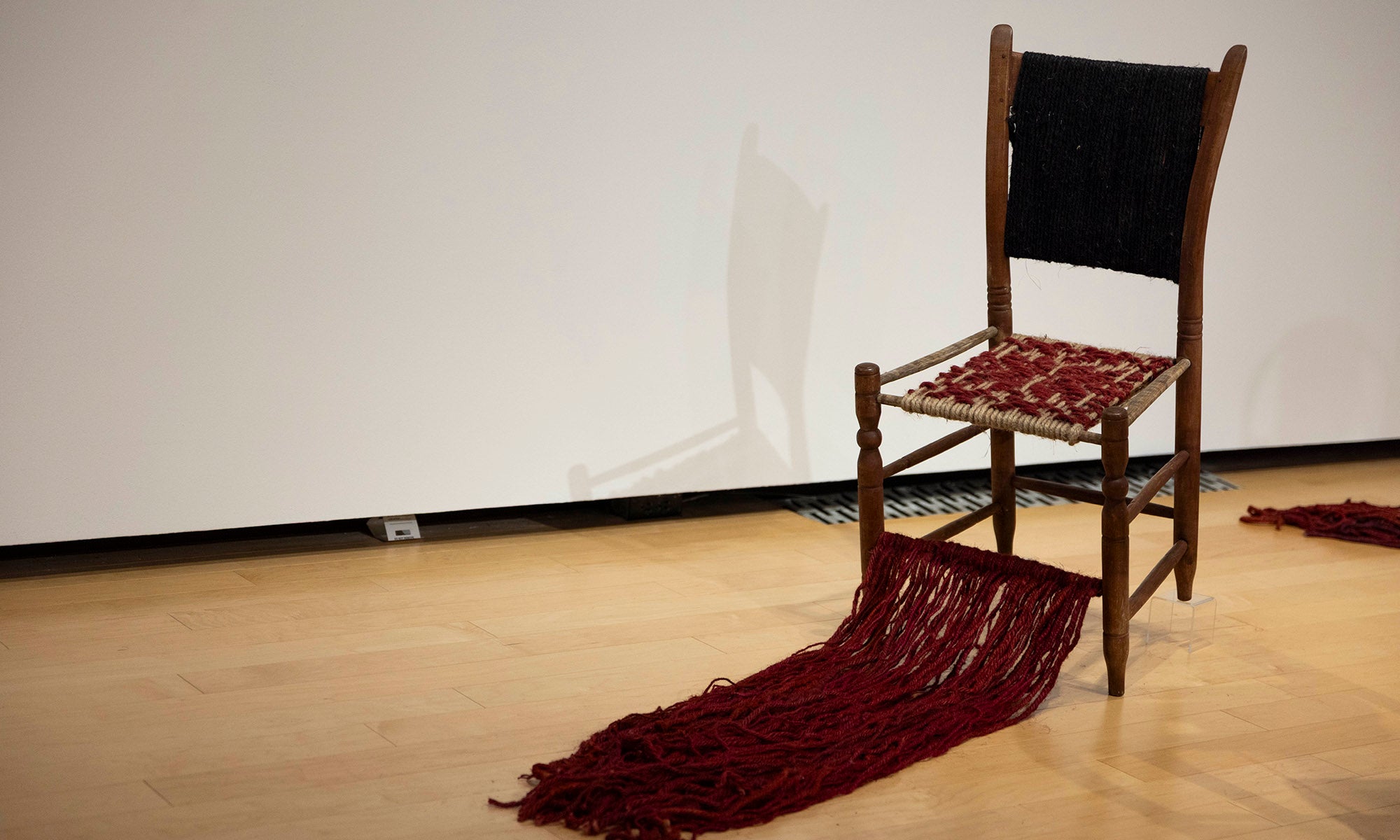 A wooden chair with a woven red seat extends its tassel-like trail onto the floor. The black fabric backrest adds contrast. Part of the "It Was Never Just Then" museum exhibition at Denison Museum, this piece stands indoors against a plain wall.