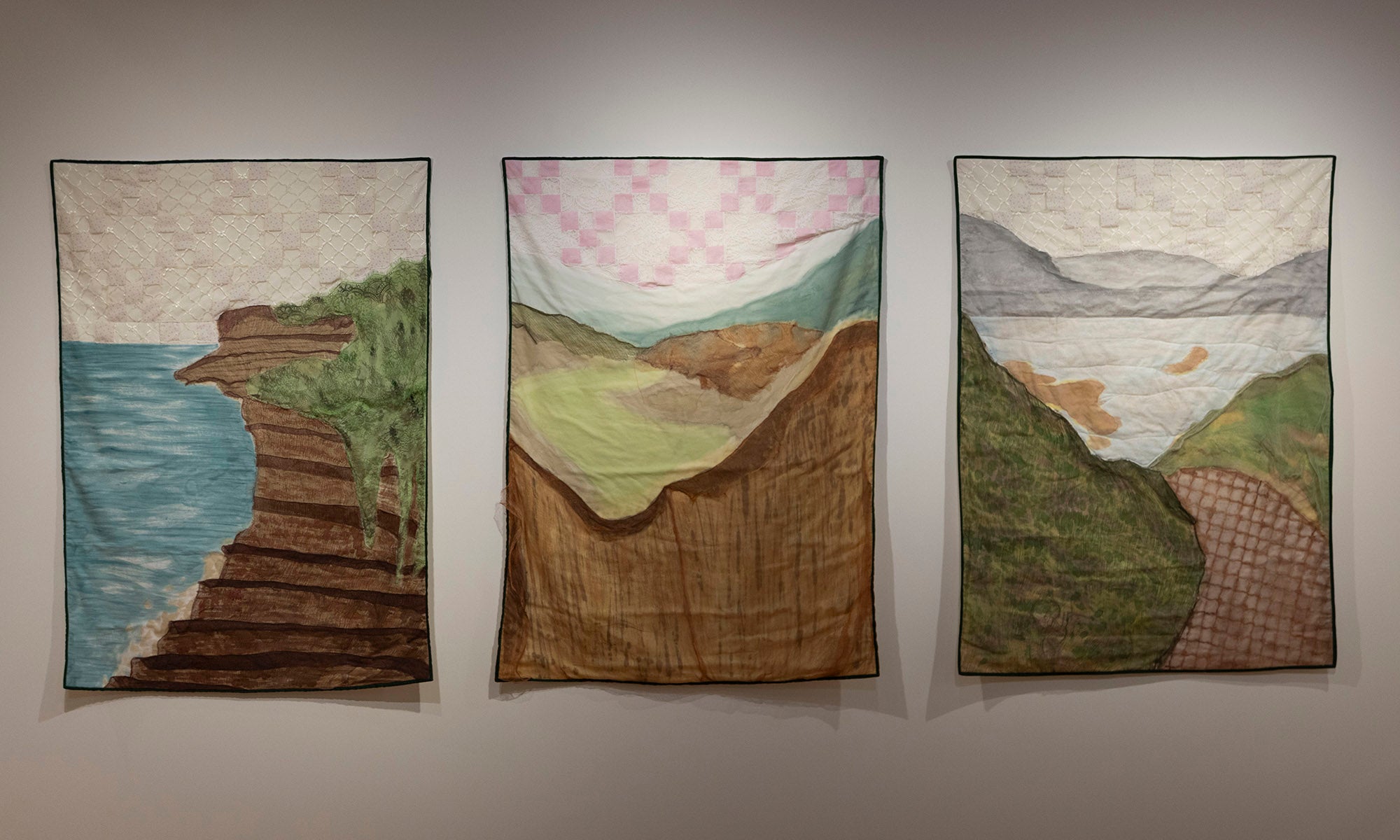 Three quilted textile artworks hang on a wall in this Denison Museum art exhibit, each depicting a different landscape scene with hills, cliffs, water, and sky in various shades of green, brown, blue, and pink.