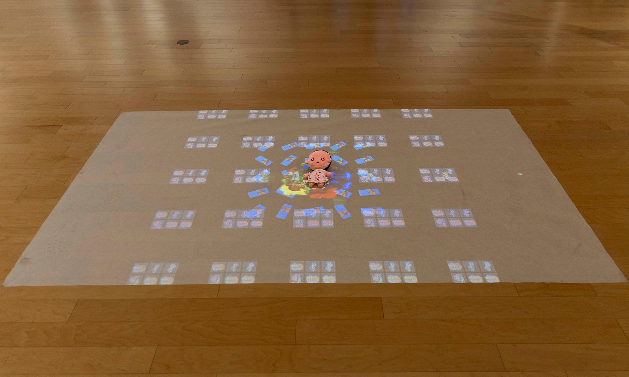 A pink toy is placed on a wooden floor at the center of a projected grid pattern with blue and white squares. Reminiscent of an art exhibition, the scene forms a rectangle around the toy, evoking themes from "It Was Never Just Then" at Denison Museum.