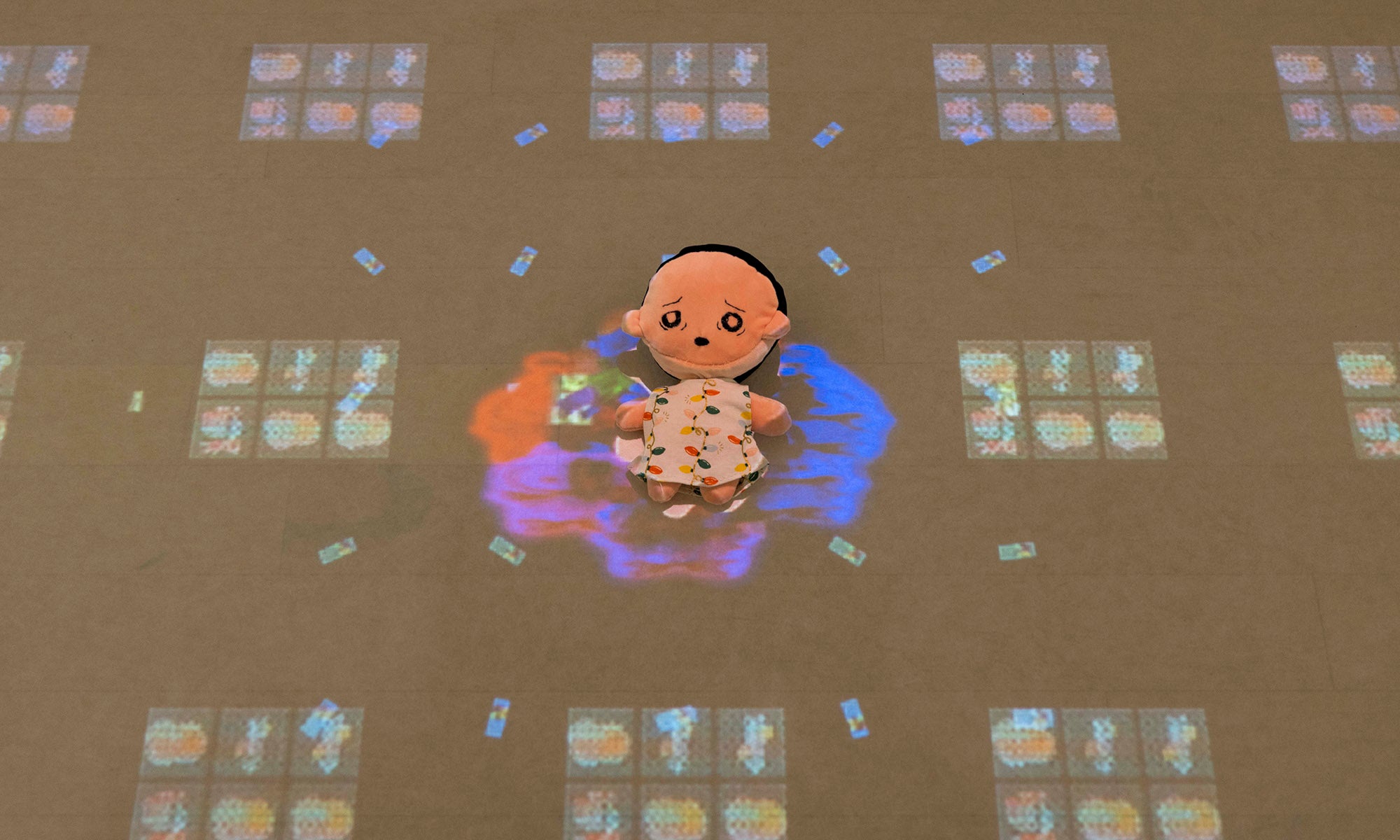 A small doll in a pink outfit sits on a surface, surrounded by colorful projected light patterns and square shapes—an enchanting scene from the "It Was Never Just Then" art exhibition at Denison Museum.