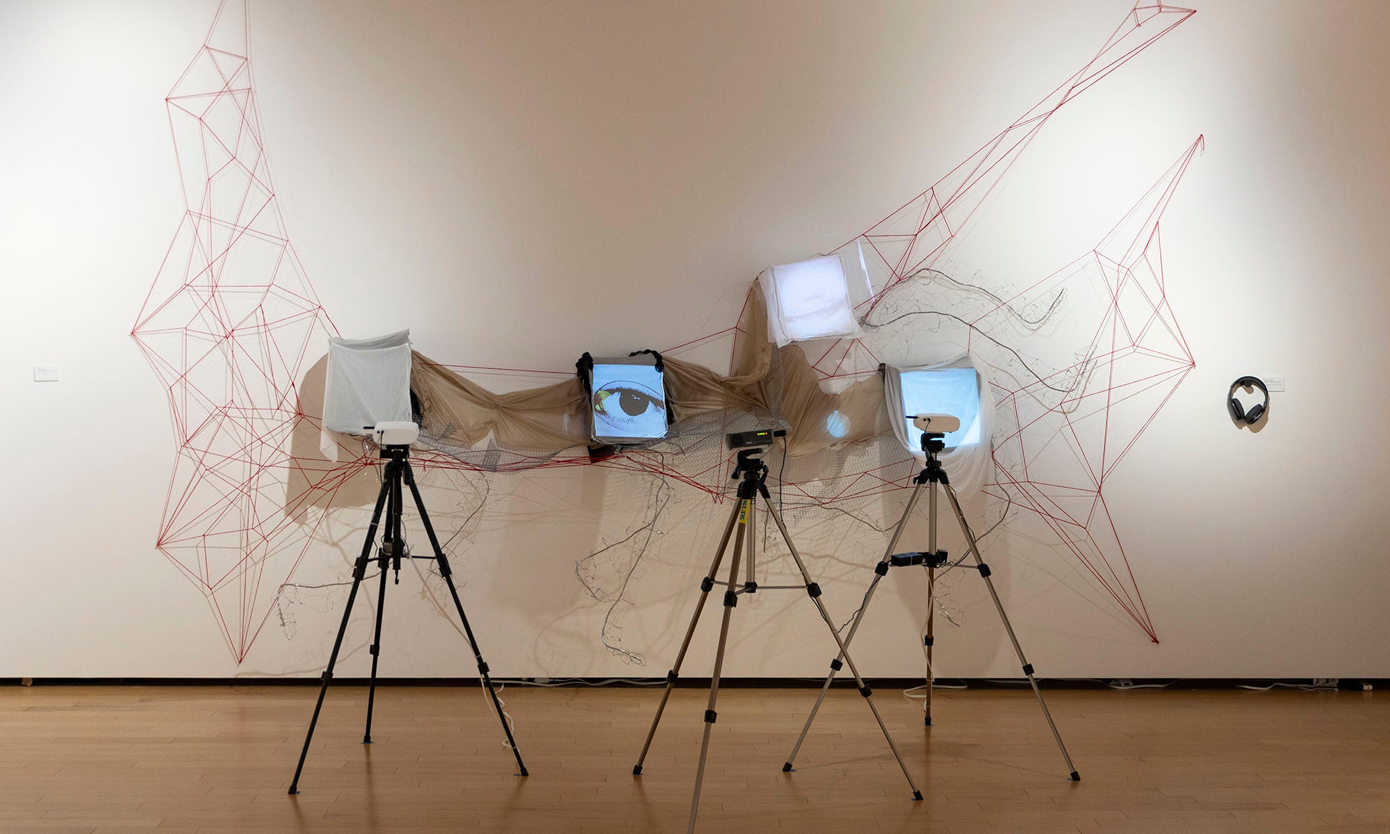 An art installation at Denison Museum’s art exhibition, titled It Was Never Just Then, features three tripods with digital screens draped in semi-transparent fabric before a white wall with a red string geometric pattern and a small circular object.