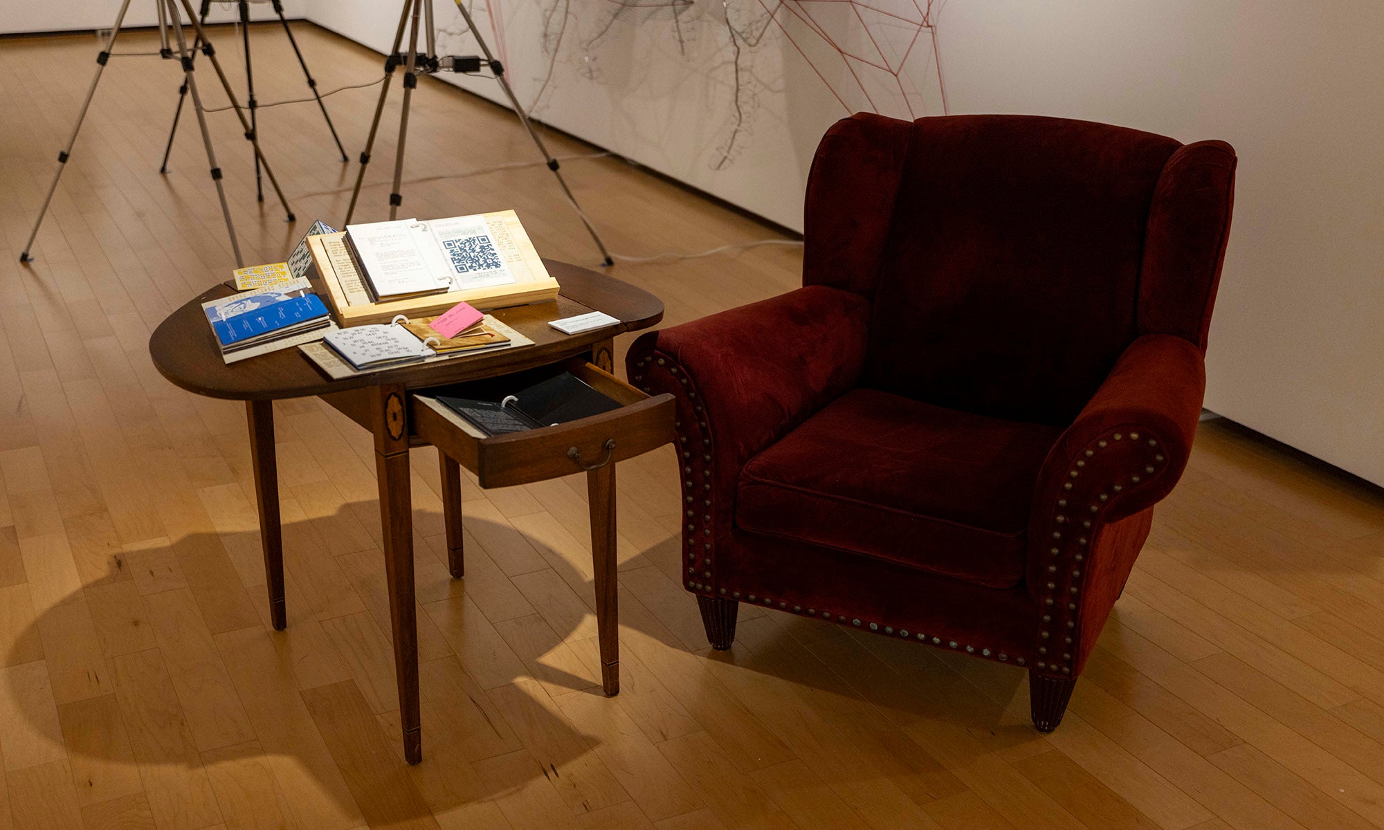 A maroon upholstered armchair sits next to a small wooden table with open books and papers in the Denison Museum gallery space, featuring wooden floors and artwork from It Was Never Just Then on the walls.
