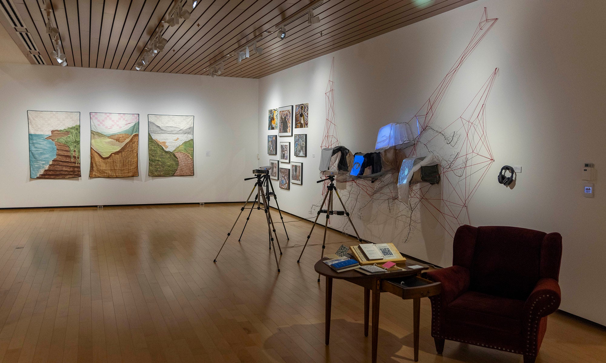 An art exhibition at Denison Museum features paintings on the walls, two cameras on tripods, a small table with open books, and a red armchair on a wooden floor under recessed lighting — inviting you into the world of "It Was Never Just Then."