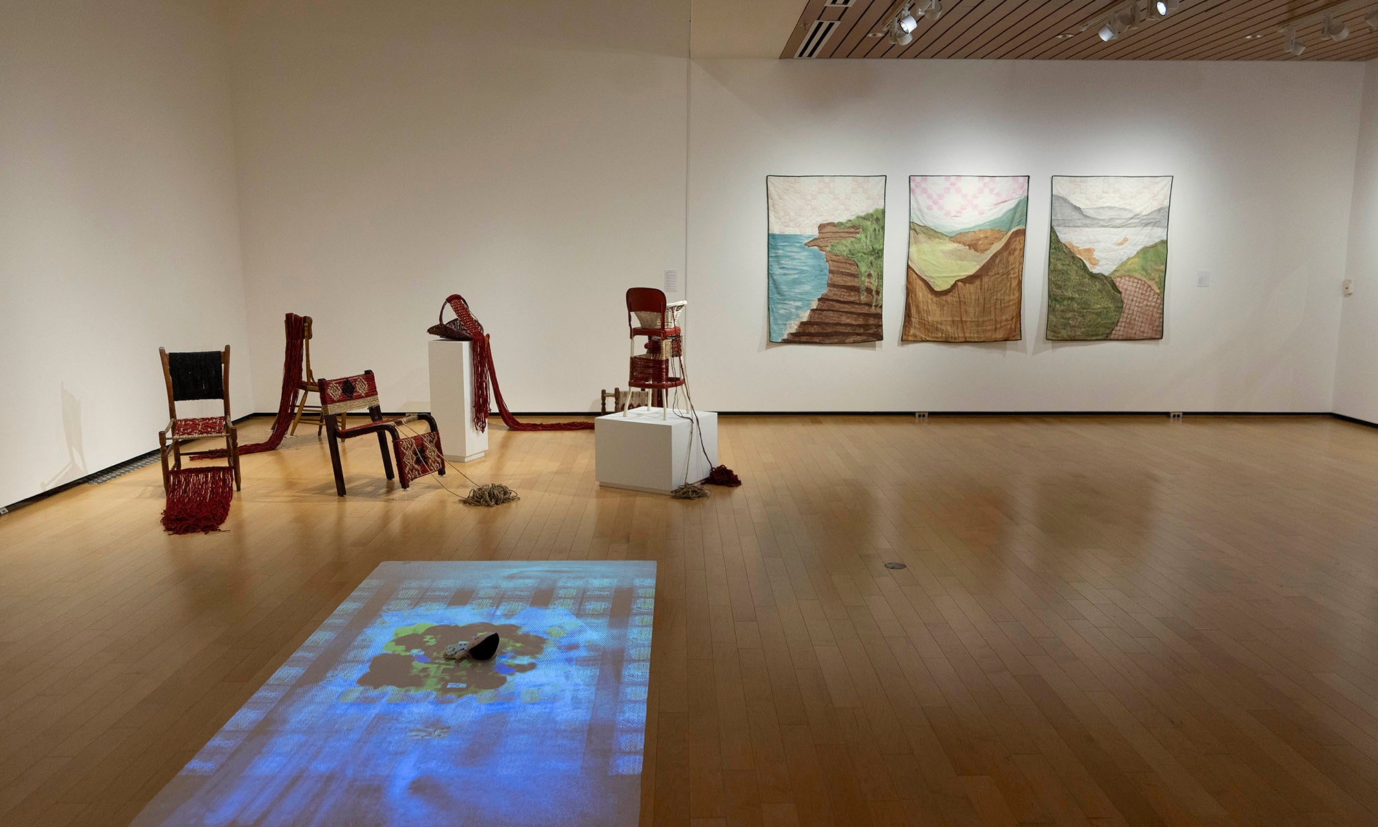 A contemporary art gallery, part of a Denison Museum exhibition, displays red chairs, baskets, and sculptures on the floor and pedestals, with three landscape paintings on the wall and a blue projection on the wooden floor.