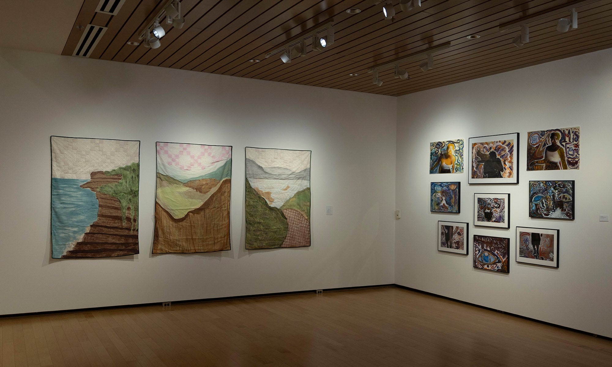 In the Denison Museum, a gallery room displays three large landscape-themed quilts on the left wall and nine smaller, framed textile artworks—including pieces from "It Was Never Just Then" — arranged in a grid on the adjacent wall under spotlights.