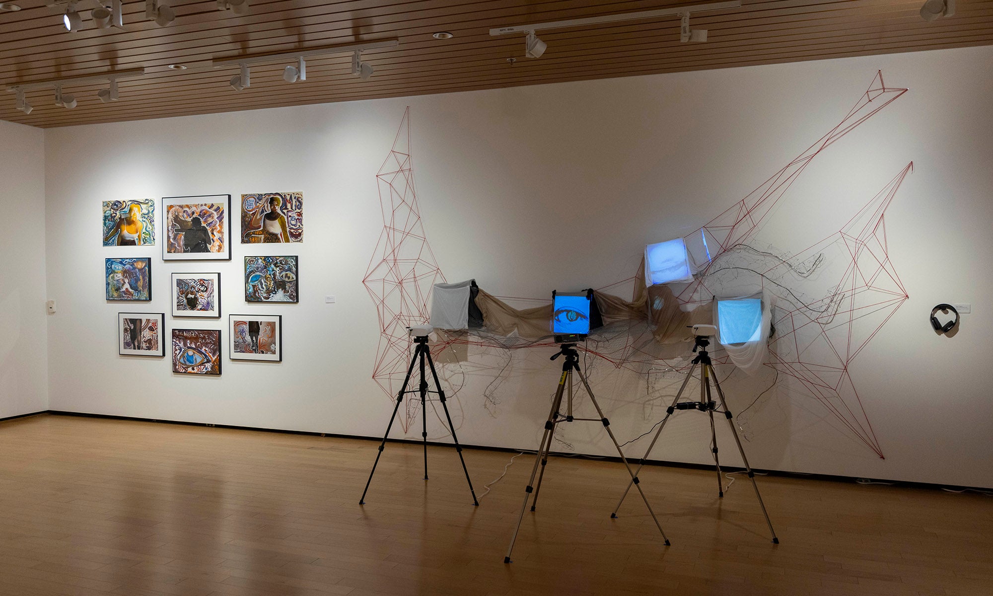 Art exhibition at Denison Museum: framed pictures line the left wall, while three monitors on tripods in the center are linked by red lines. A pair of headphone hangs on the wall to the right.