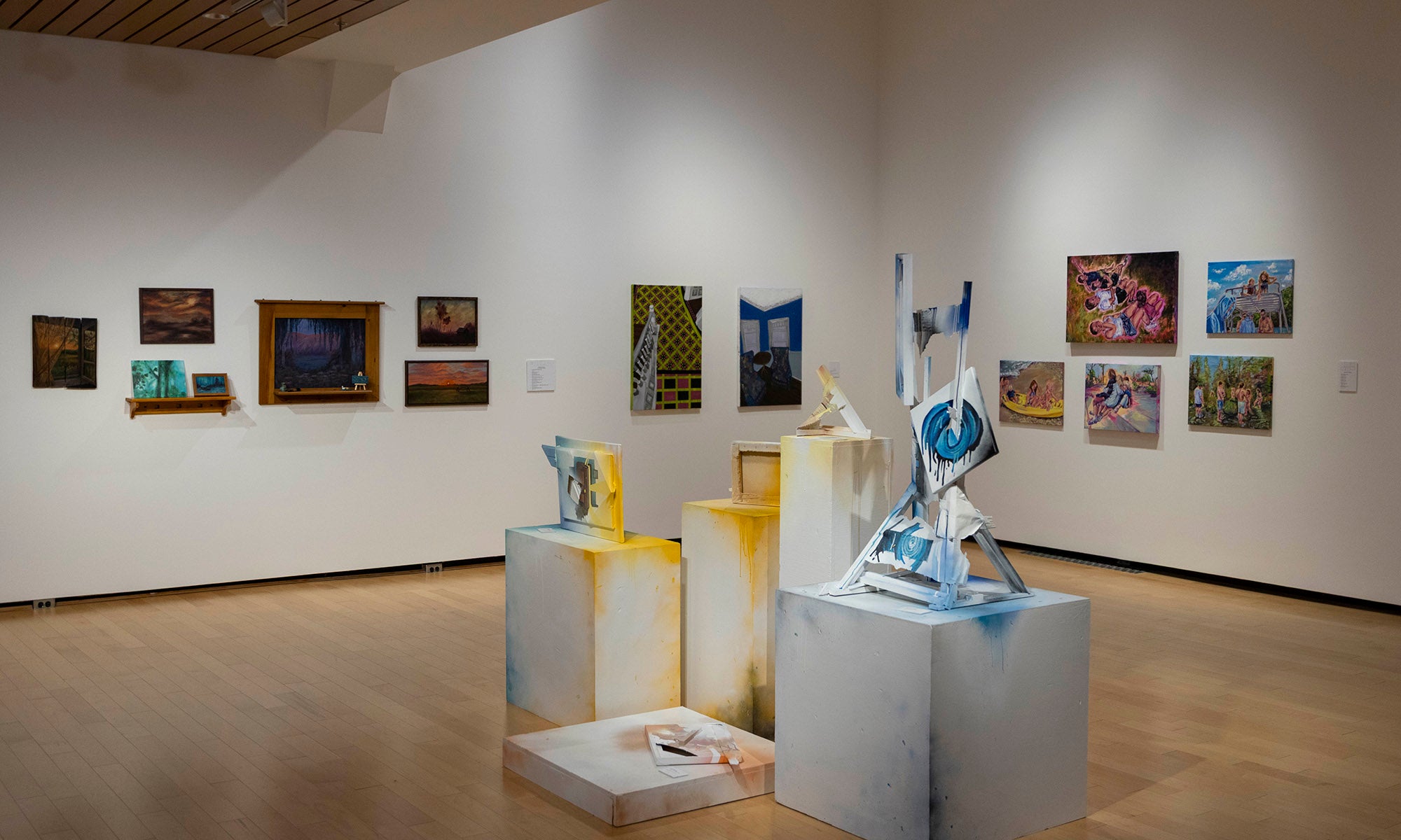 An art exhibition at Denison Museum features hardwood floors, colorful paintings on white walls, and several abstract sculptures on white pedestals in the center of the room.
