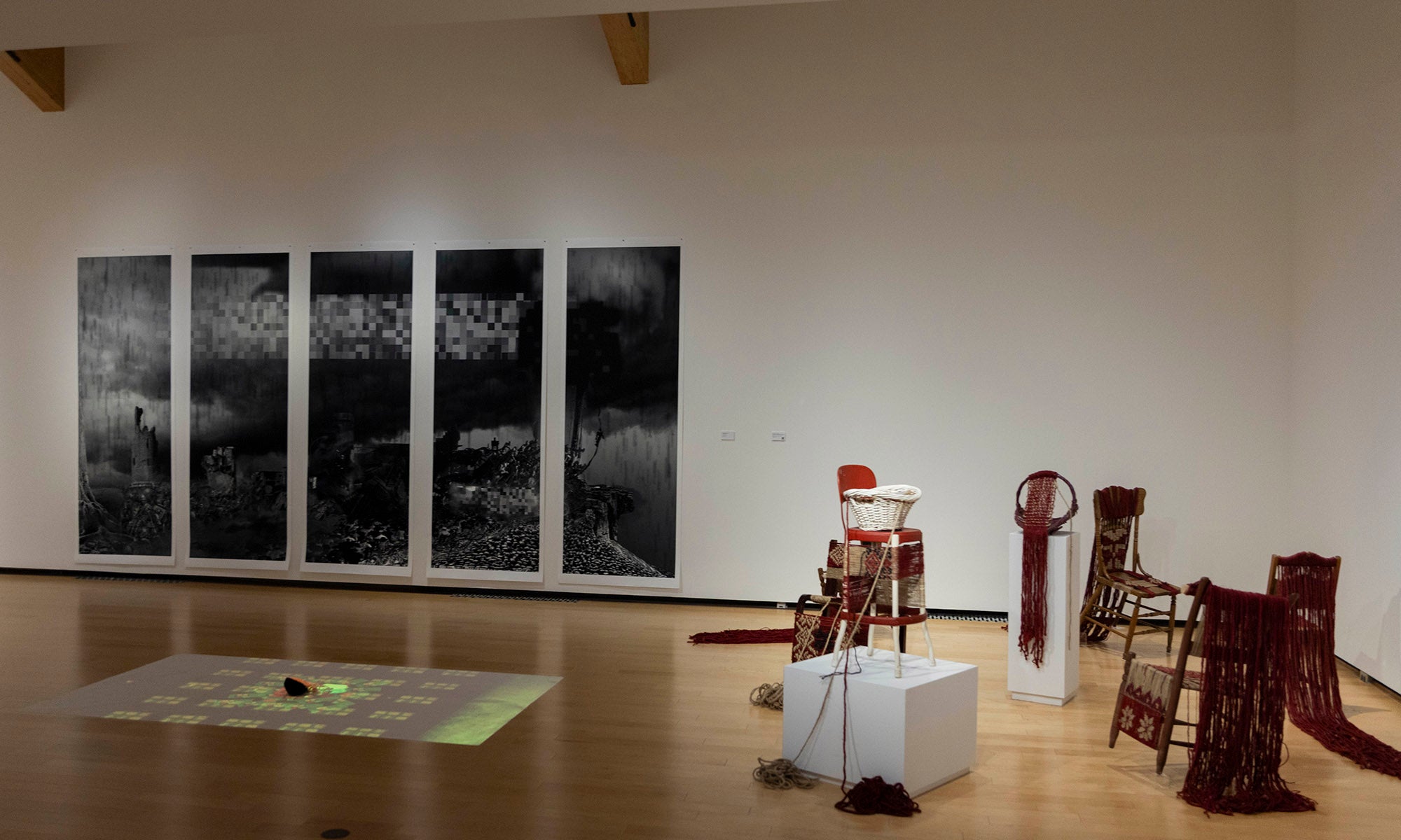 A gallery space at Denison Museum features five large black-and-white photographic panels on the wall and a mixed-media installation of chairs, fabric, pedestals, and objects as part of the art exhibition "It Was Never Just Then."