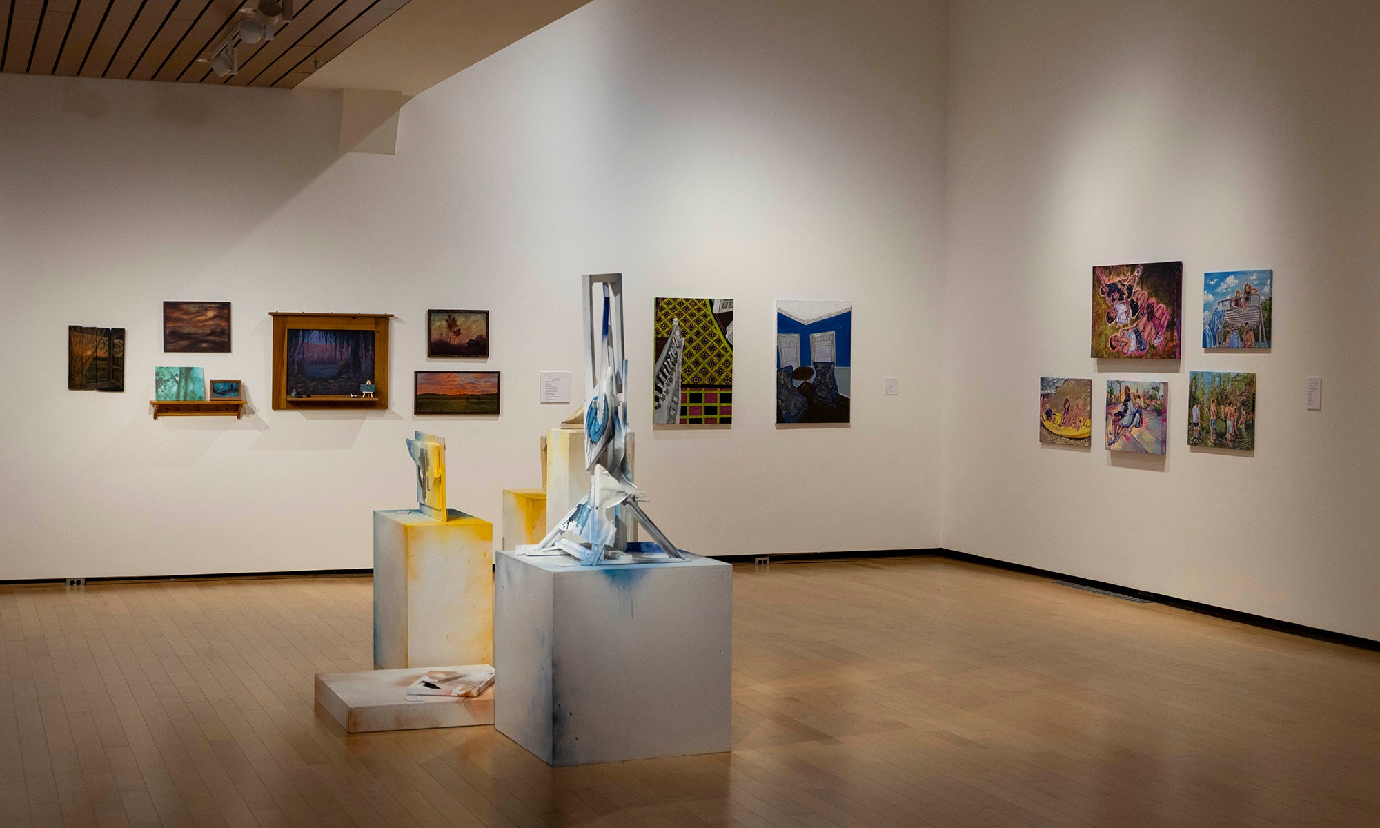 A contemporary art gallery reminiscent of a museum exhibition, with paintings and photographs on white walls and several abstract sculptures displayed on the floor — evoking the spirit of Denison Museum’s “It Was Never Just Then.”