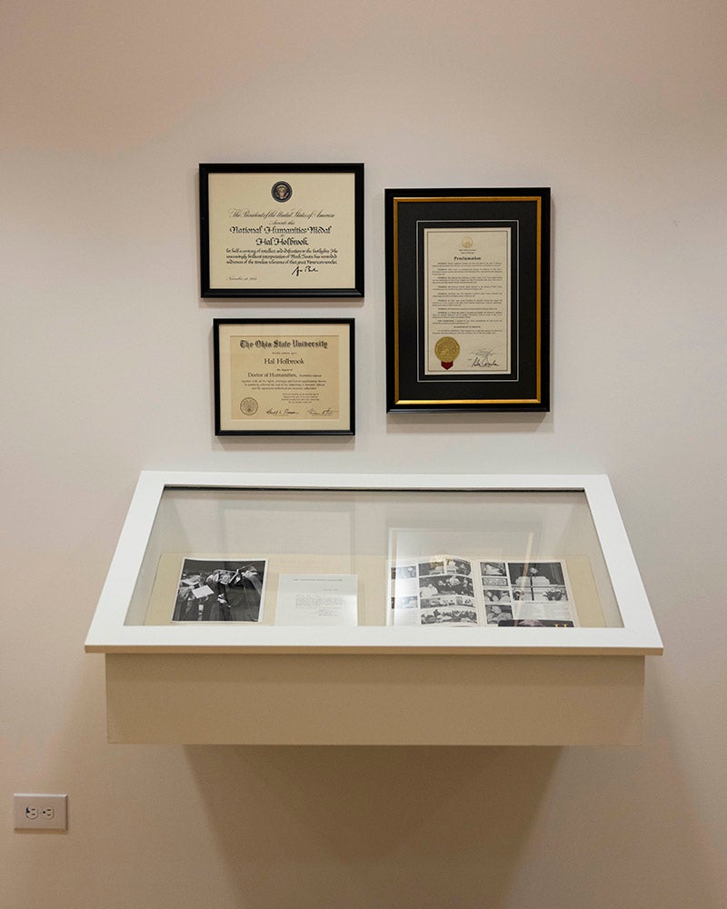 A display case with documents and photos sits below three framed certificates or diplomas on a plain, light-colored wall at the Denison Museum. The case contains open books and papers arranged neatly for viewing.