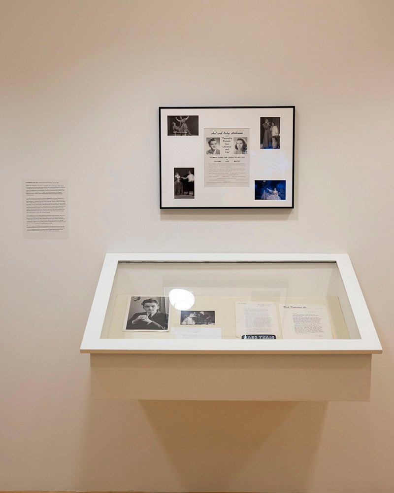 A museum display at Denison Museum features documents and photos in a glass case below a framed informational panel with text and images, all mounted on a plain beige wall near Broadway.