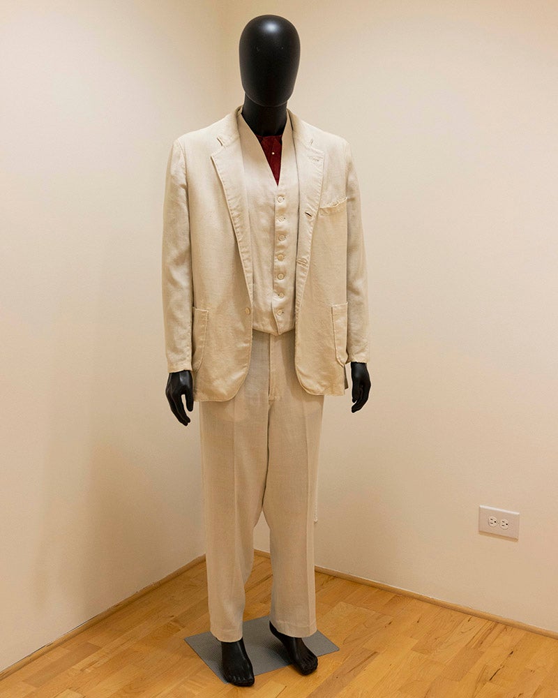 A mannequin dressed in a cream-colored three-piece suit with a matching jacket, vest, and trousers stands in a sparsely decorated room at the Denison Museum, with light walls and a wooden floor.