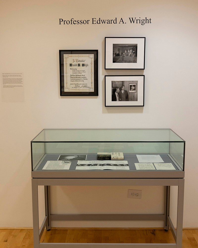 A glass display case with documents and photographs beneath it sits against a white wall labeled Professor Edward A. Wright in the Denison Museum. Three framed items, including photos and a certificate, are mounted on the wall above the case.