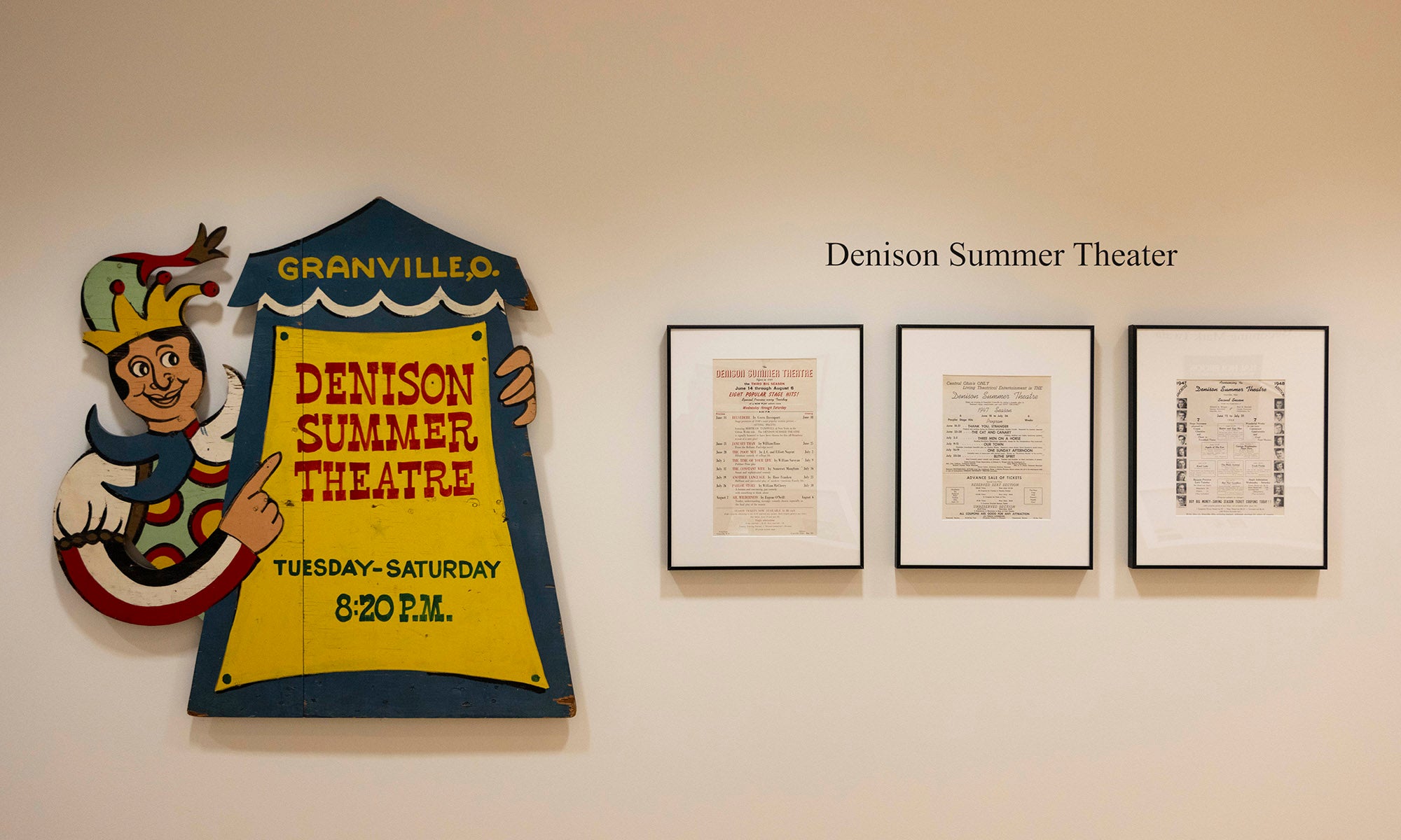 A vintage Denison Summer Theatre sign shaped like a clown stands next to three framed documents displayed on a beige wall under a sign that reads Denison Summer Theater, evoking the charm of Broadway and the history of the Denison Museum.