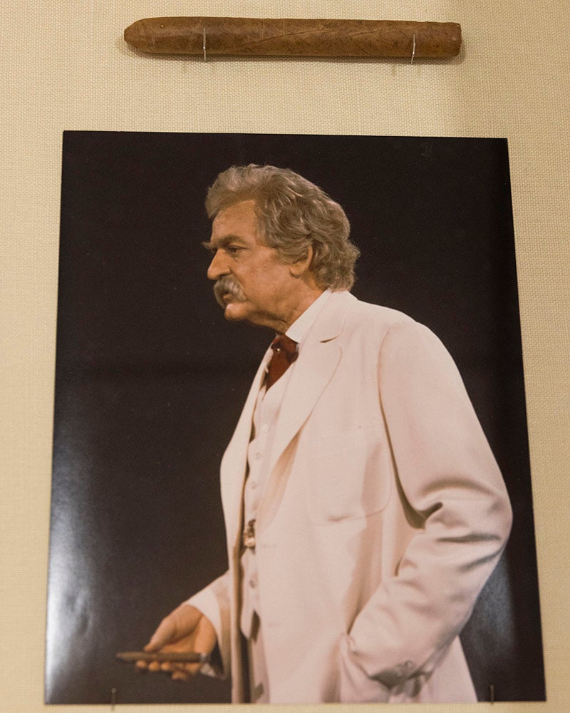 A framed photograph from the Denison Museum shows an older man with gray hair and a mustache in a white suit, holding an object in his right hand. Above the photo, a cigar is mounted horizontally on the wall.