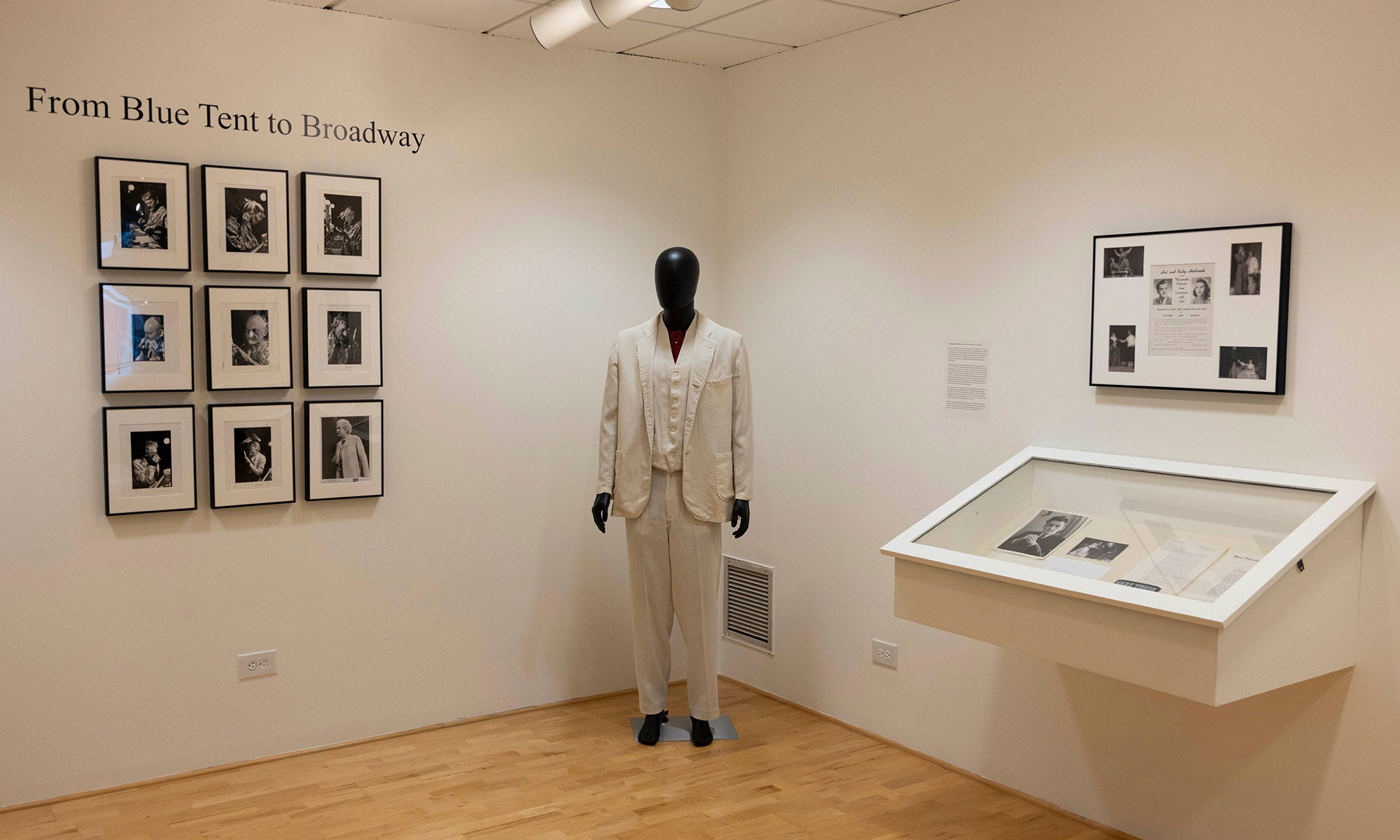 An art gallery corner at Denison Museum featuring a wall display of nine framed photographs, a mannequin dressed in a light-colored suit, a framed collage on the right wall, and a glass display case with documents and images from Broadway.