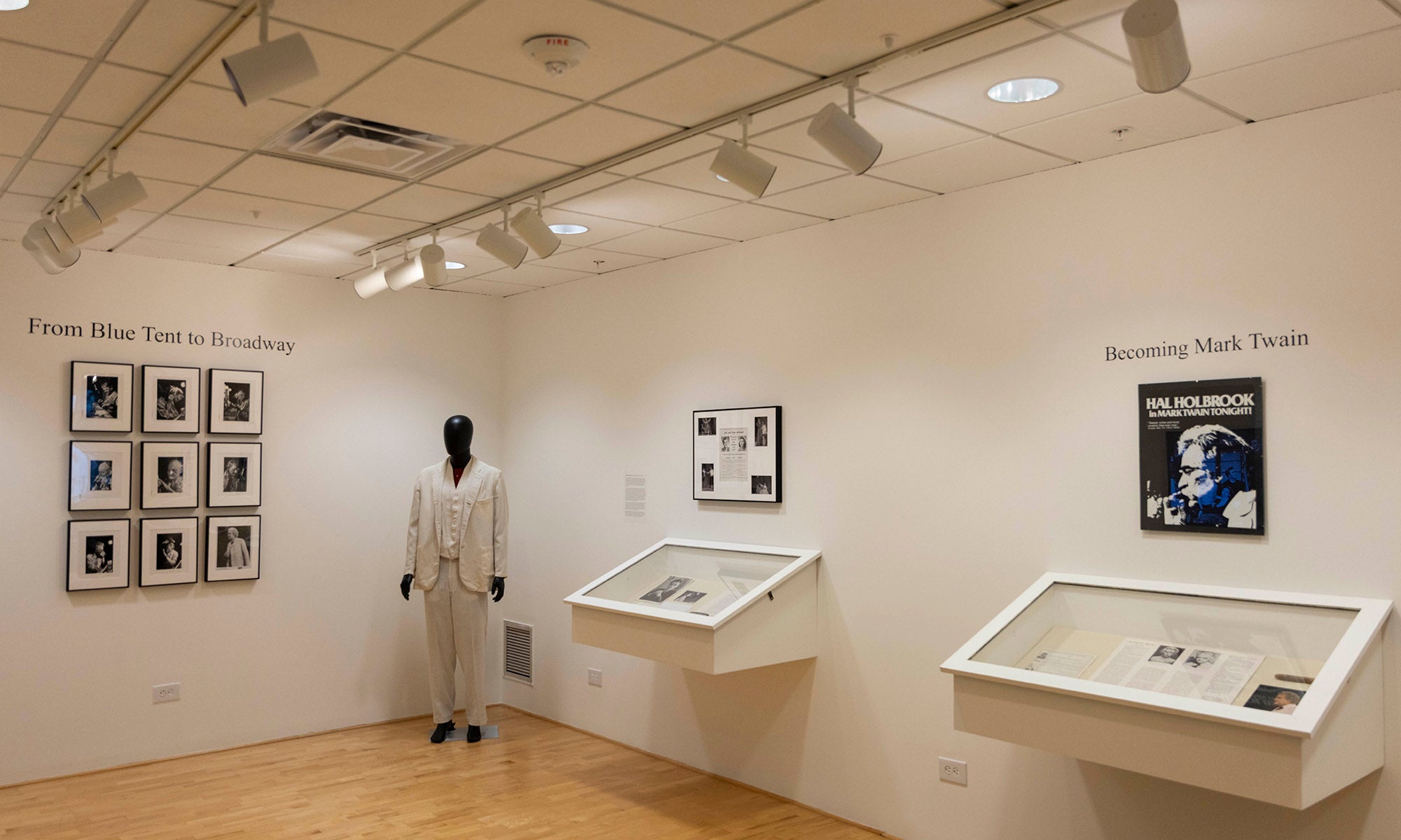 A gallery room at the Denison Museum with white walls displays framed photographs, a mannequin dressed in white clothing, and two glass exhibit cases. Track lights illuminate the exhibits, and text is visible on the walls above the displays.