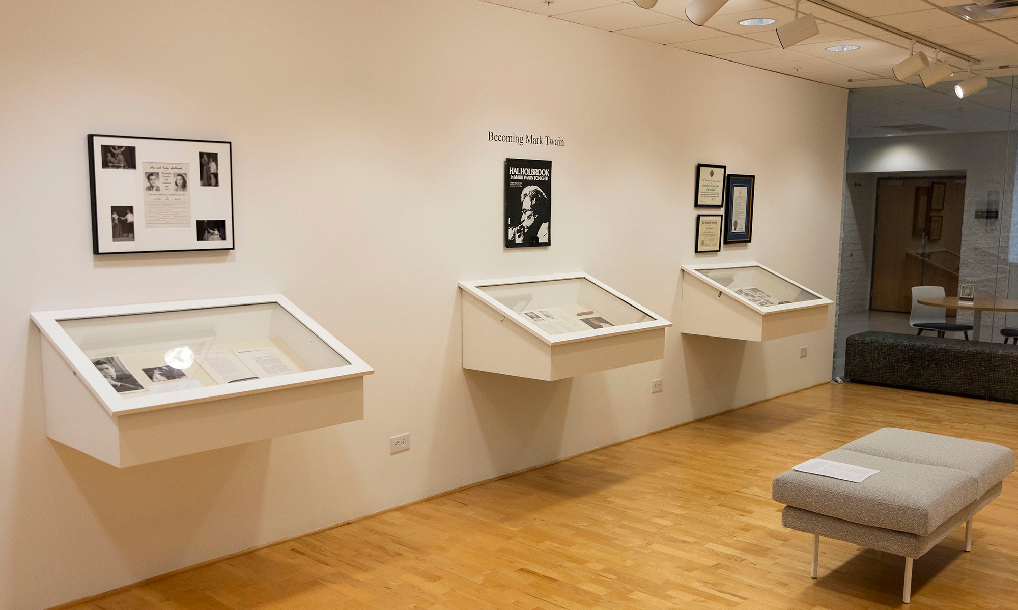 A museum exhibit room at Denison Museum with three wall-mounted display cases containing documents and artifacts, framed pictures and text panels above each case, a bench in the foreground on a wooden floor, inspired by the Blue Tent on Broadway.