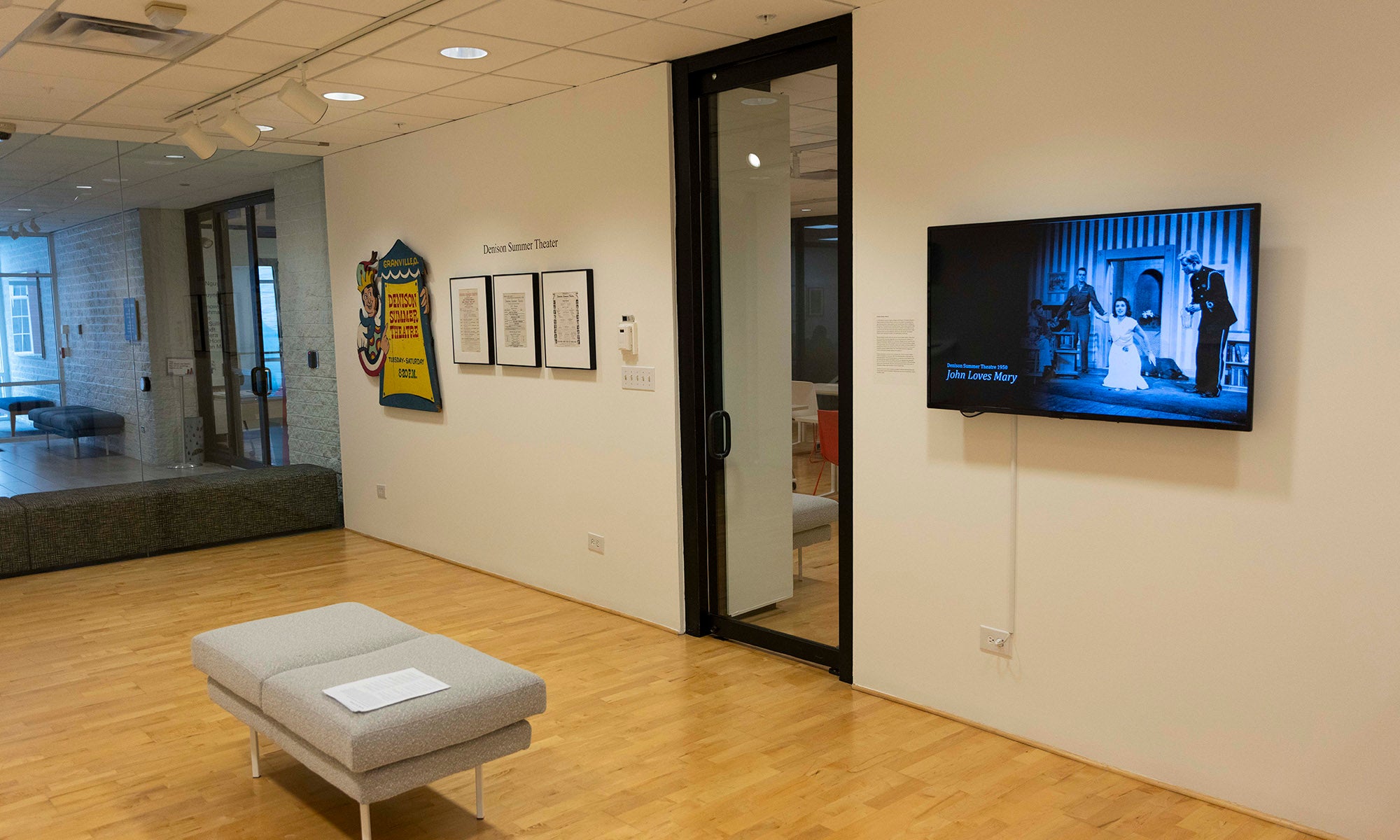 A modern art gallery room at Denison Museum with wooden floors, a bench in the center, framed artworks on the wall, a glass door, and a digital screen displaying an image of people in a blue-toned scene.