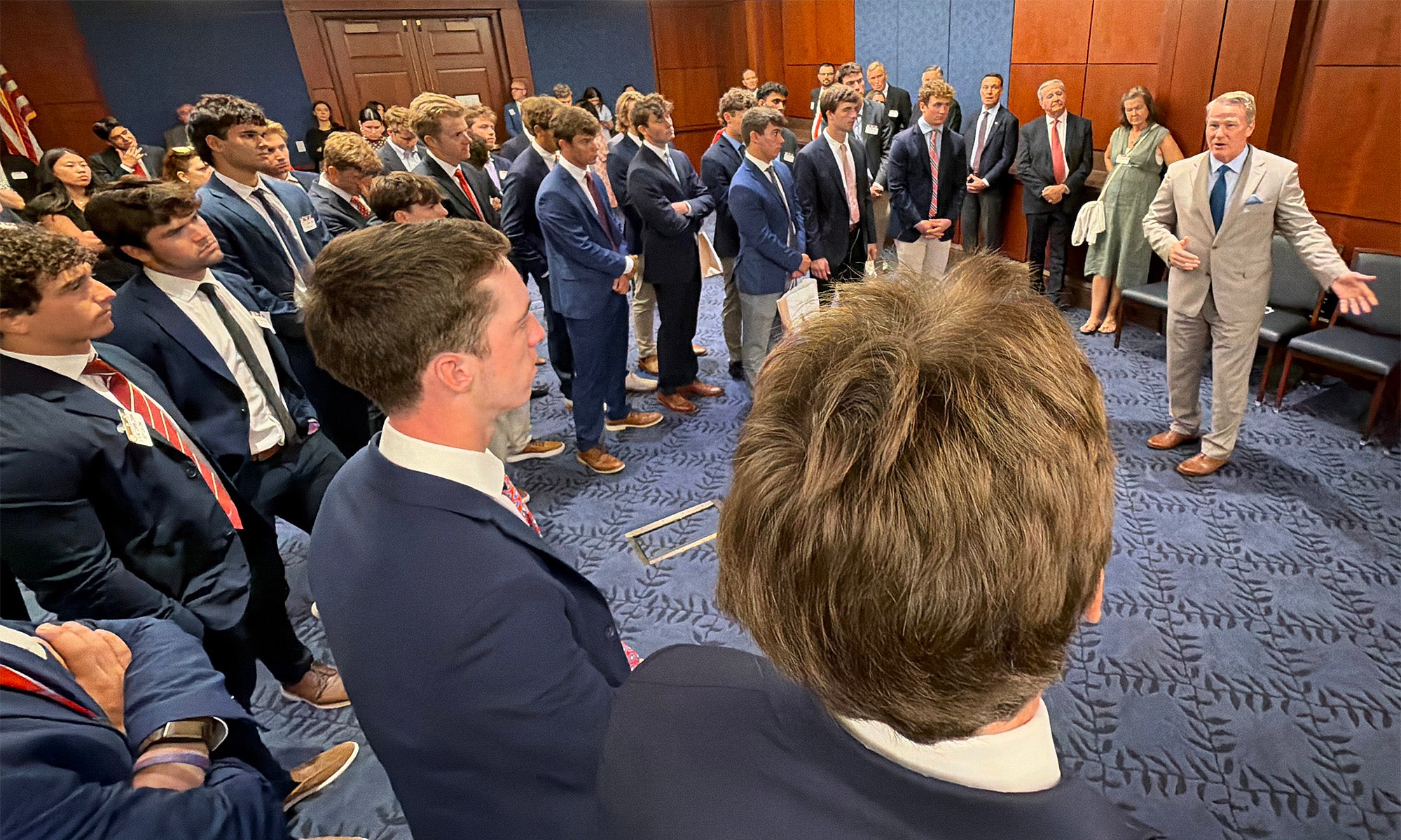 A group of men in suits, including several distinguished Big Red alumni, listen to a speaker in a formal room with wooden walls and blue carpeting.