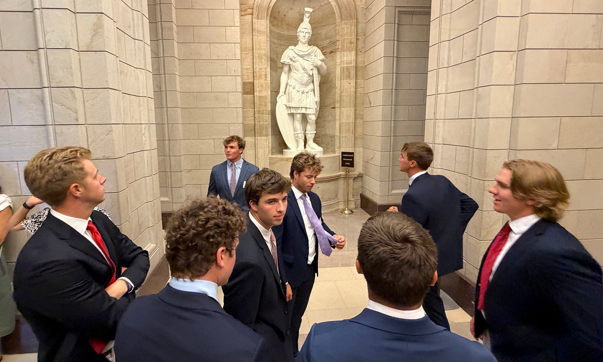 A group of men in suits stand and talk in a stone hallway with a large statue in the background.