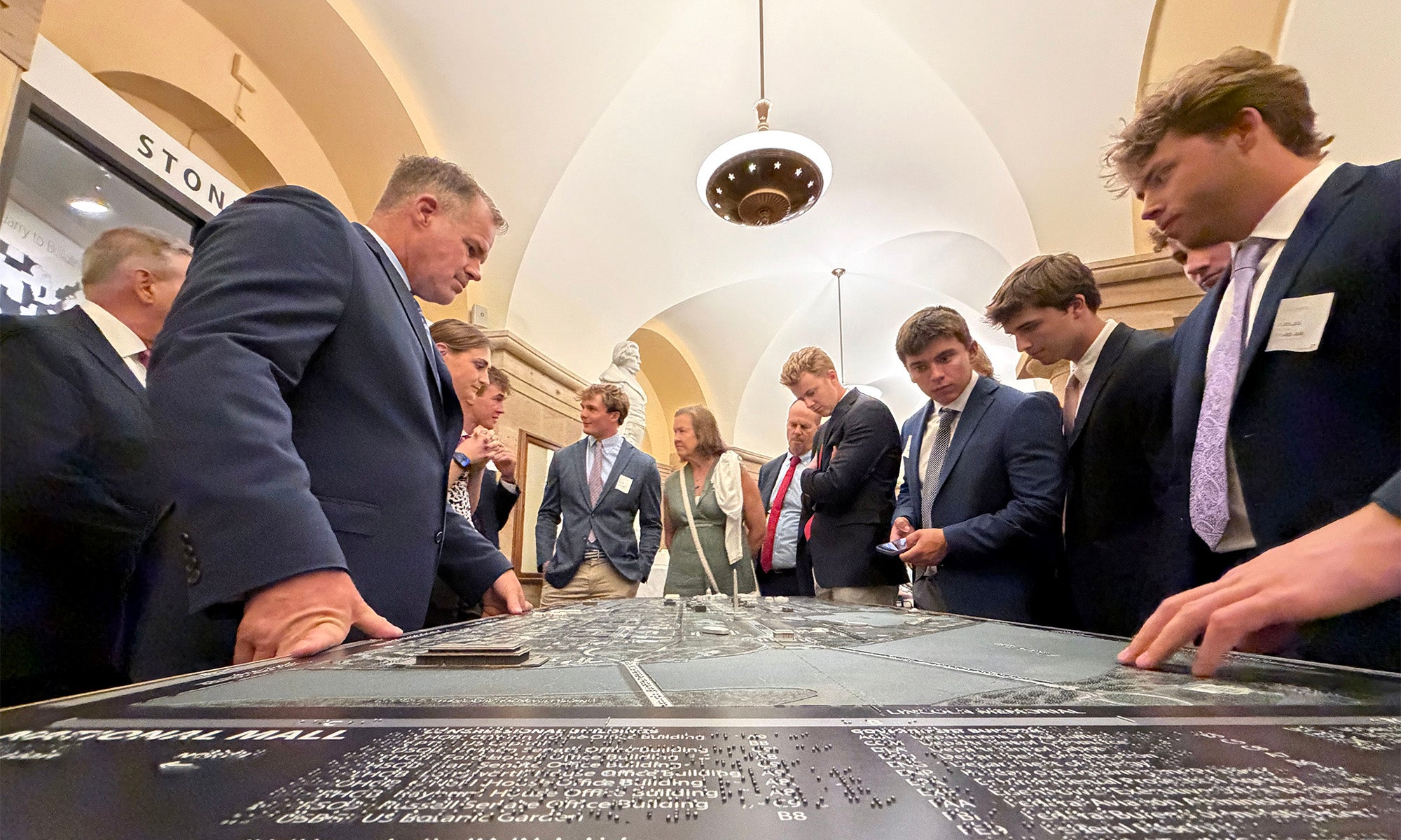 A group of people in suits stand around a large table with a map or display, engaged in discussion inside a well-lit room with arched ceilings.