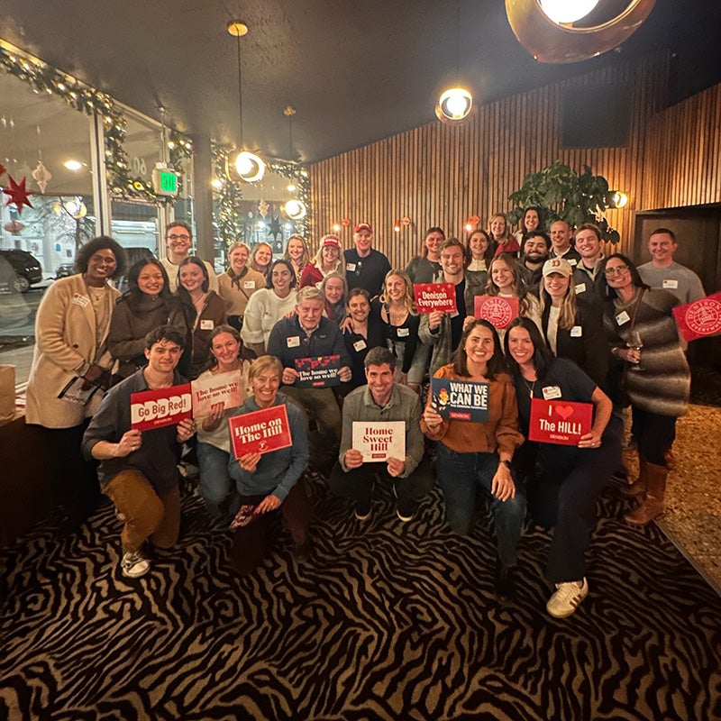 A group of about 35 people pose indoors on a zebra-print carpet, some holding signs with text. There are string lights and wood-paneled walls in the background. The group appears to be at a social event or gathering.